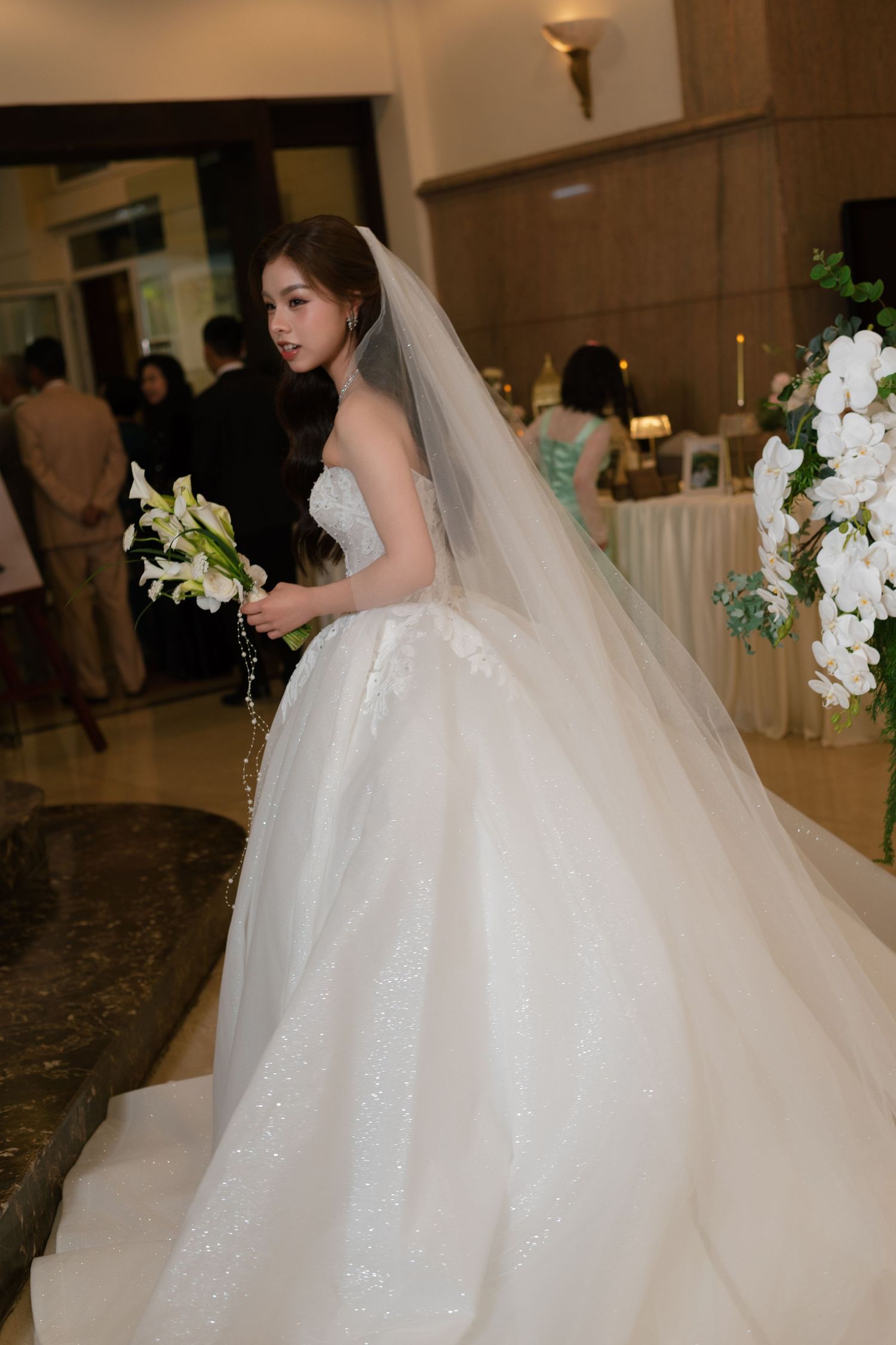 A bride in a stunning white ballgown with a long veil holds white flowers at an elegant wedding reception venue.