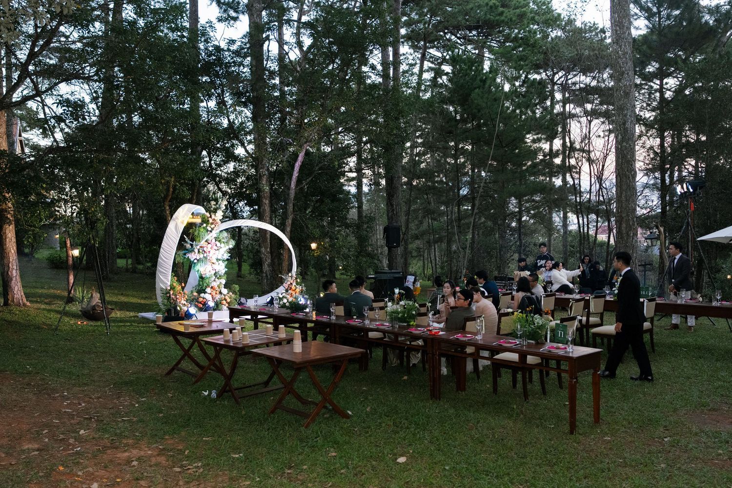 Elegant outdoor wedding reception setup with long wooden tables, floral arch, and string lights in a pine forest at dusk.