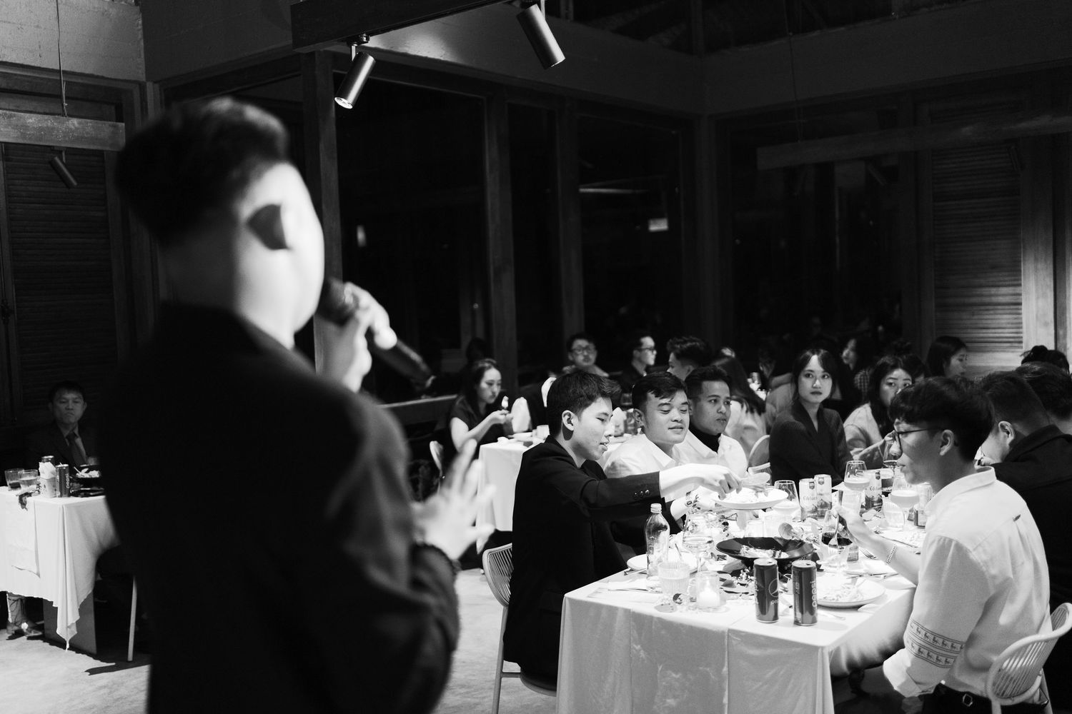 Black and white photo of a groom addressing seated wedding reception guests at elegantly set dinner tables.