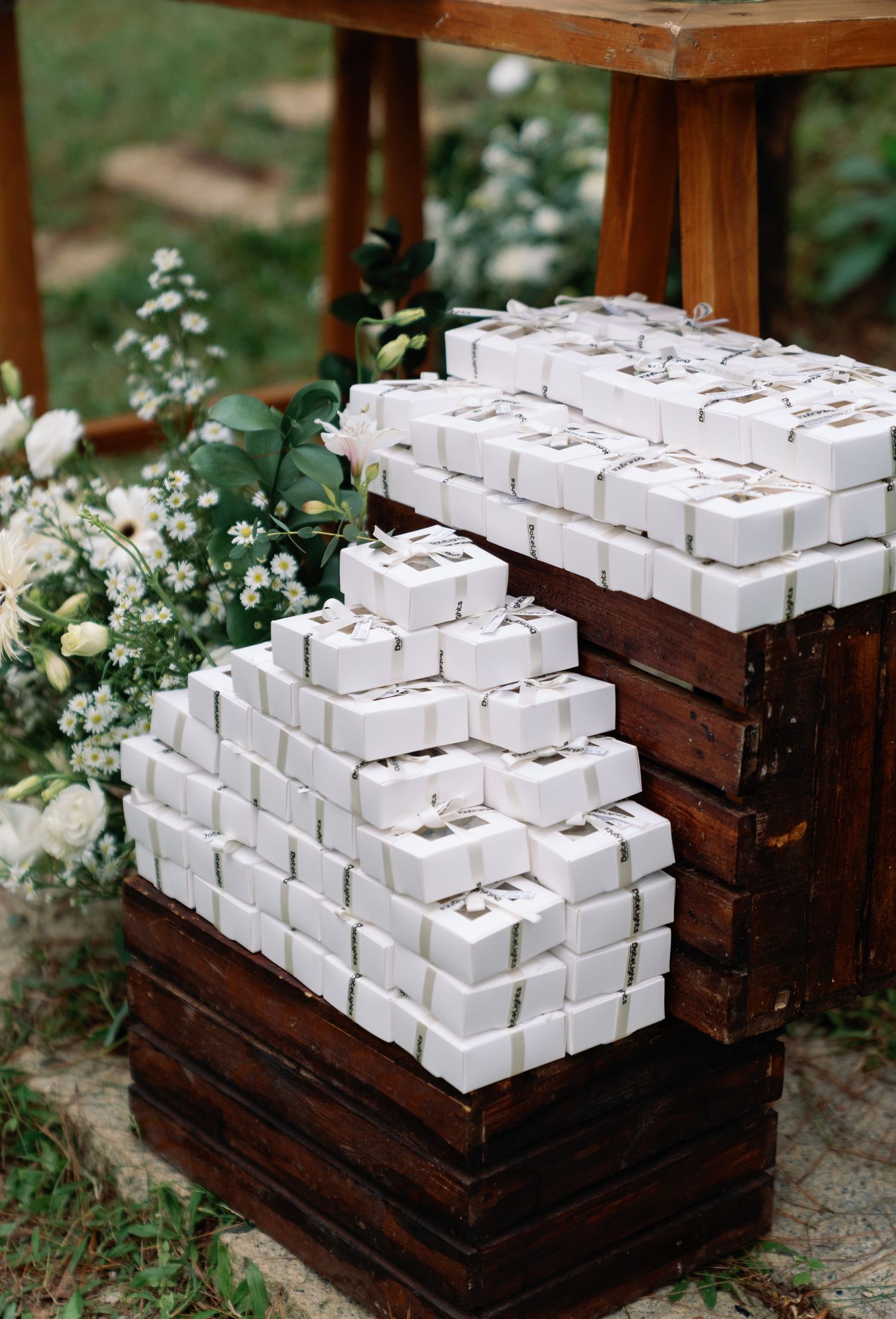 White wedding favor boxes stacked on rustic wooden crates surrounded by white floral arrangements.