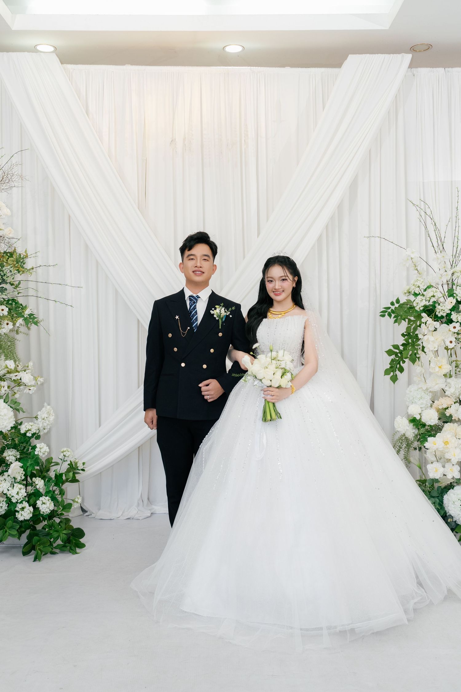 Vietnamese bride and groom posing before elegant white draped wedding backdrop with floral arrangements.