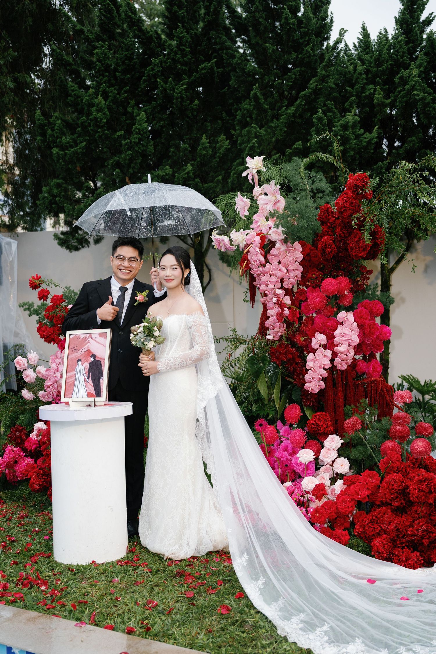 Bride and groom pose under clear umbrella at outdoor wedding ceremony with red and pink floral backdrop.