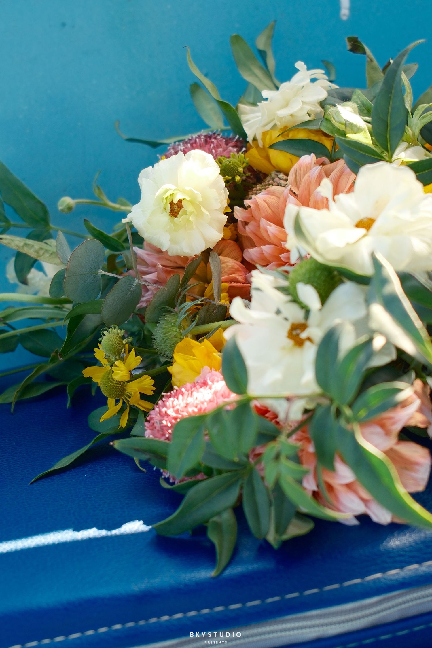 Colorful wedding bouquet with white peonies, peach dahlias, and yellow wildflowers resting on a bright blue surface.