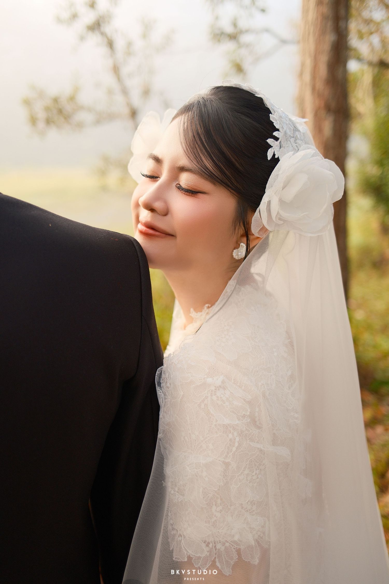 Bride in white lace wedding dress and veil leans against groom with eyes closed in serene outdoor portrait.