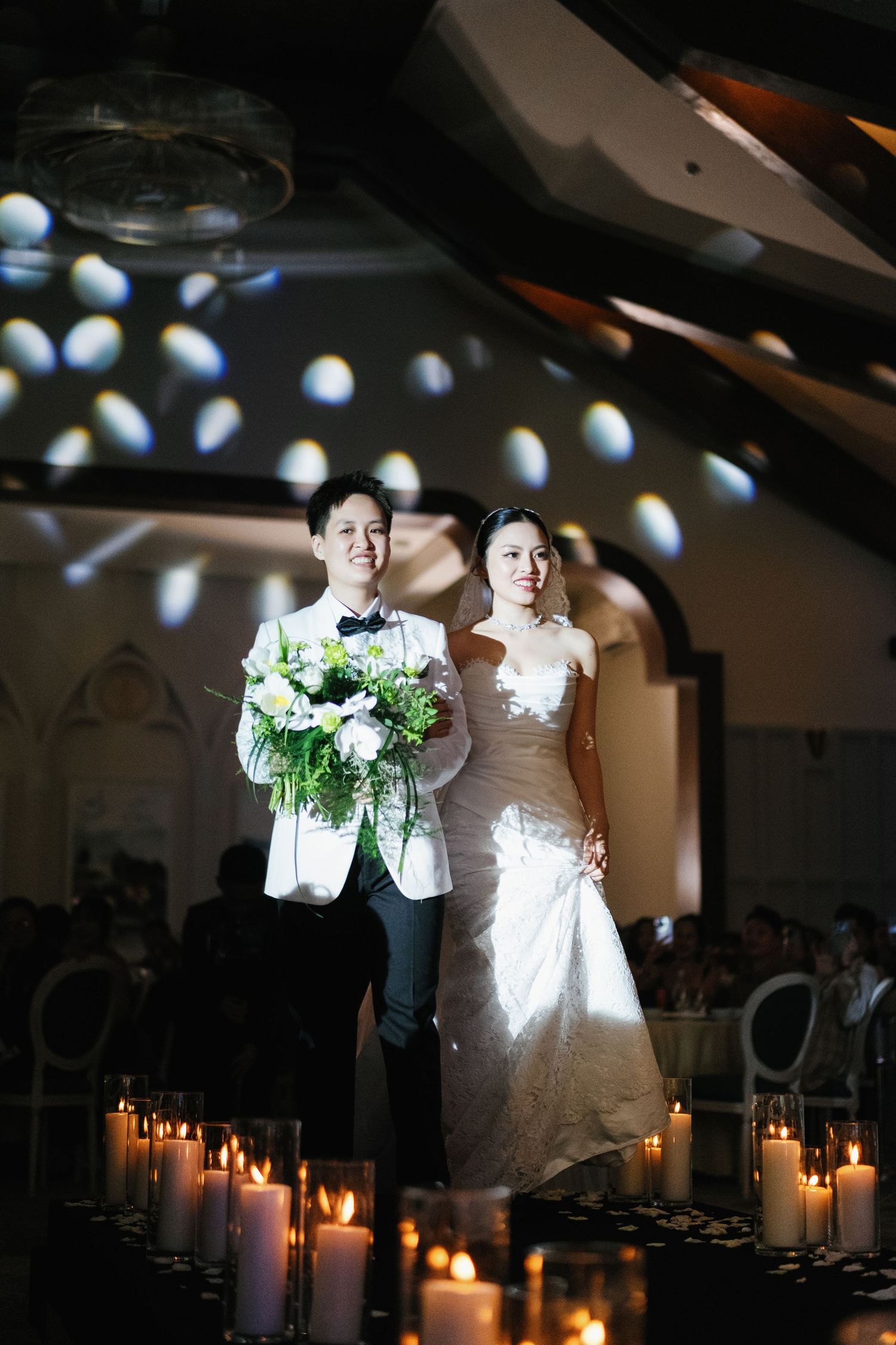 Smiling couple walking down candlelit wedding aisle, groom in white tuxedo holding floral bouquet.