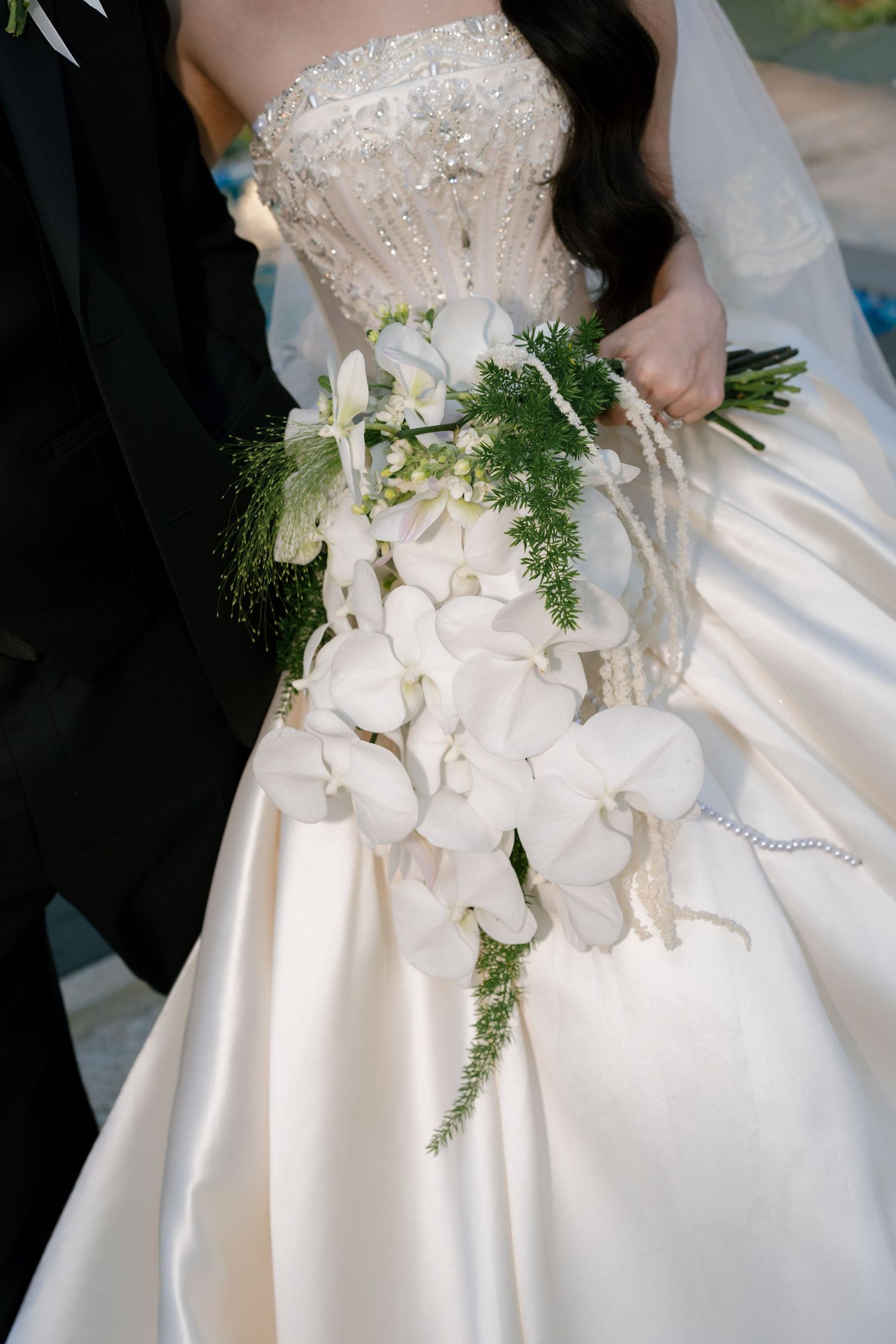 Bride in a strapless beaded wedding gown holding a cascading white orchid bouquet with greenery beside a groom in a black suit.