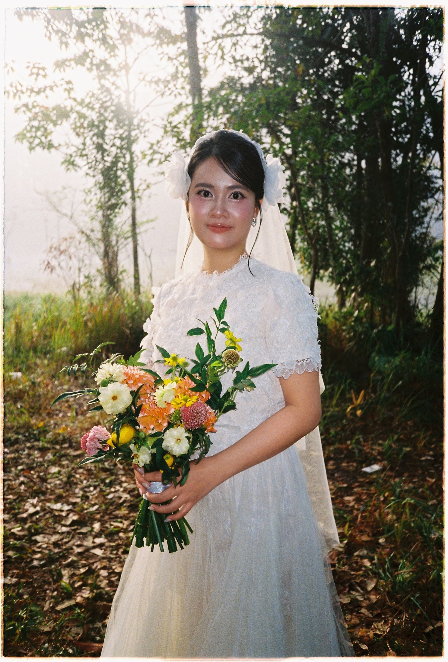 Smiling bride in white lace dress and veil holds vibrant bouquet among autumn trees in sunlight.