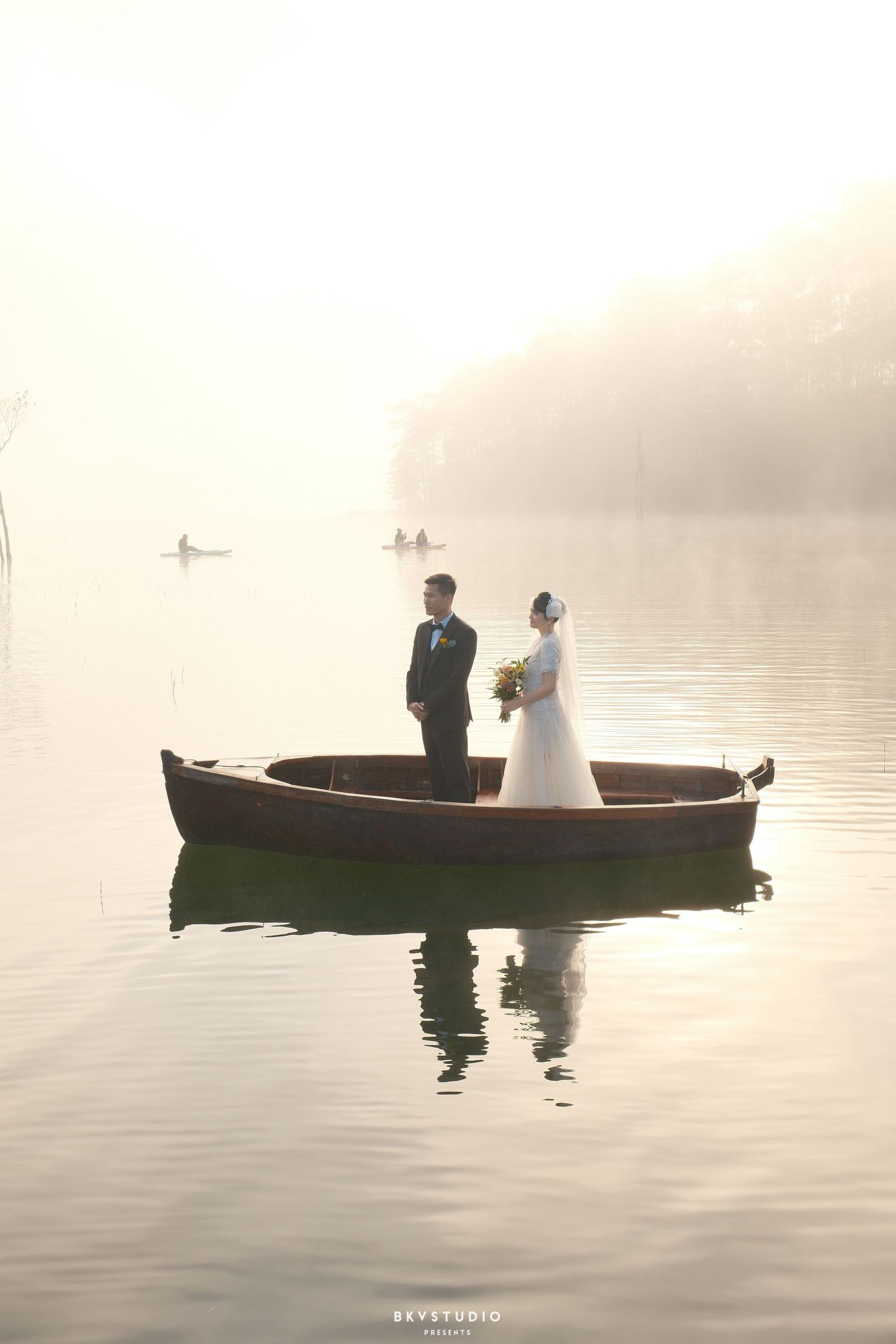 A bride and groom stand in a wooden boat on a misty, glassy lake at sunrise.