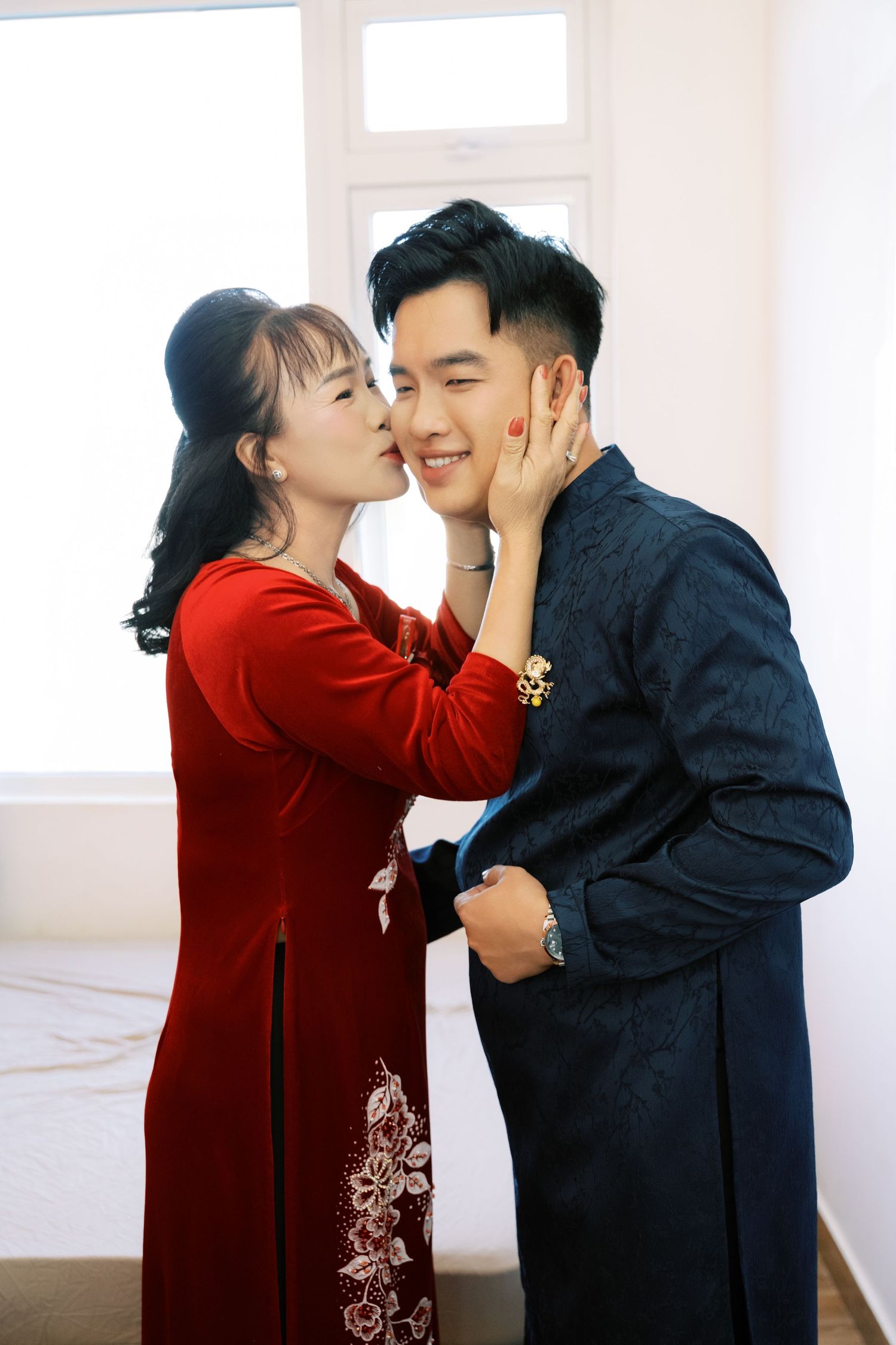 Vietnamese couple in traditional ao dai attire sharing a tender moment, woman in red kissing mans cheek.