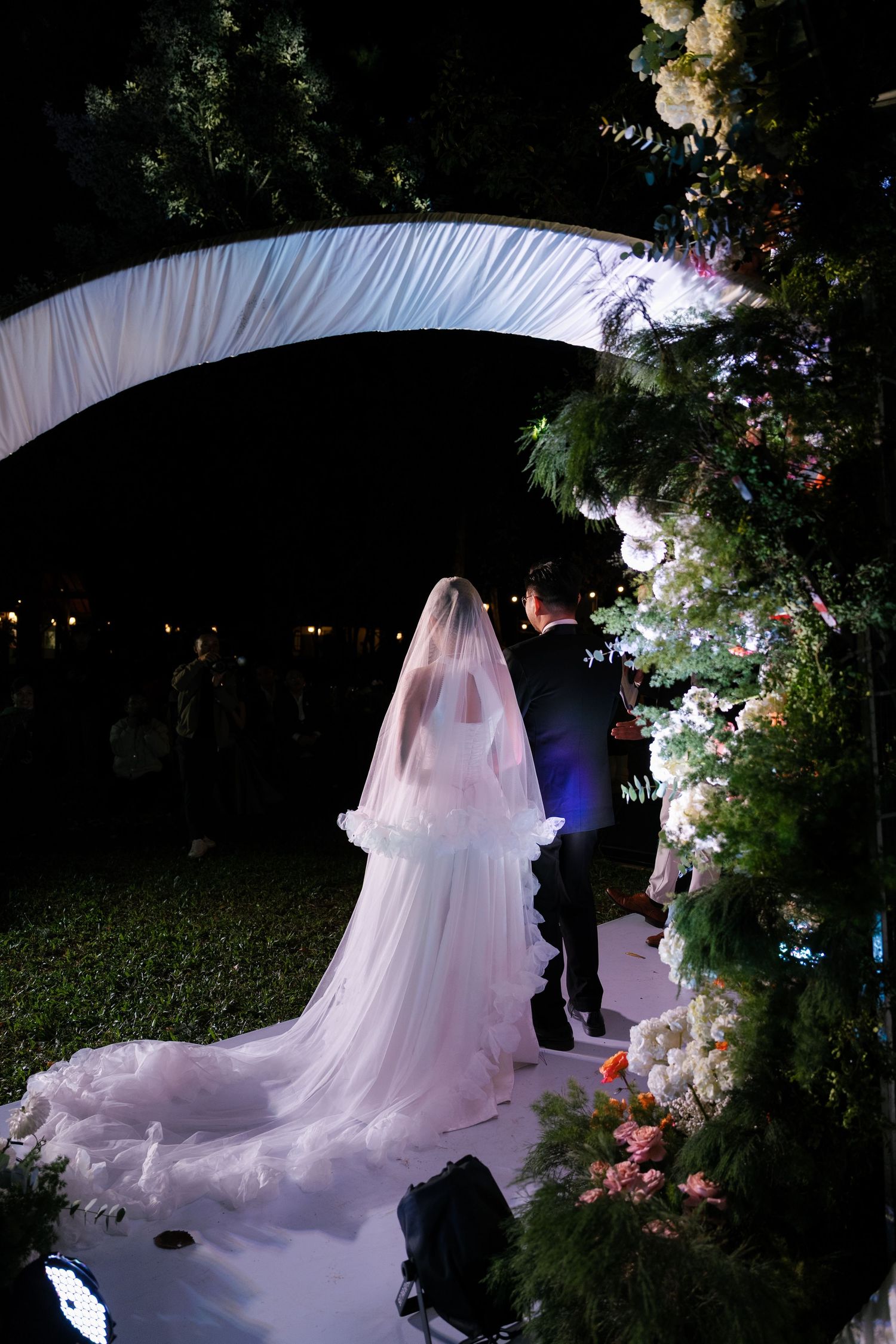 Bride with long cathedral veil walks with groom under white arch at outdoor evening wedding.