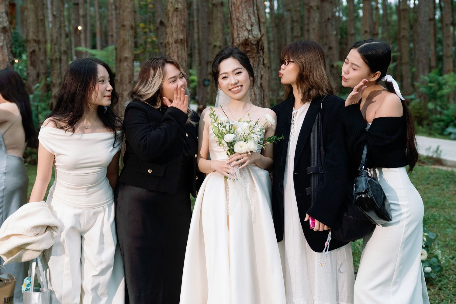 Bride in white gown holds bouquet surrounded by smiling bridesmaids in white and black outfits in a forest setting.