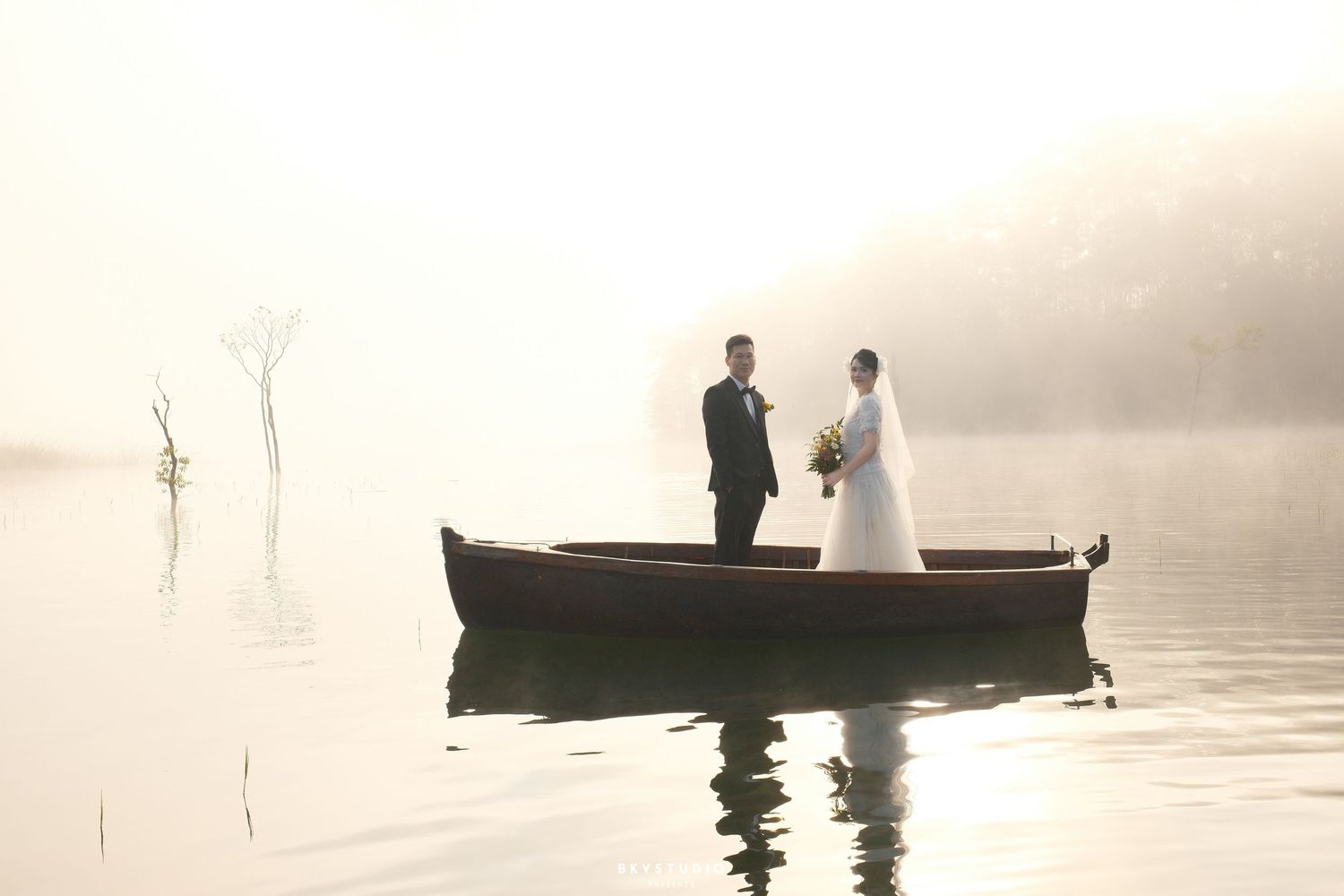 Bride and groom standing in a wooden boat on a misty lake during a romantic outdoor wedding photo shoot.