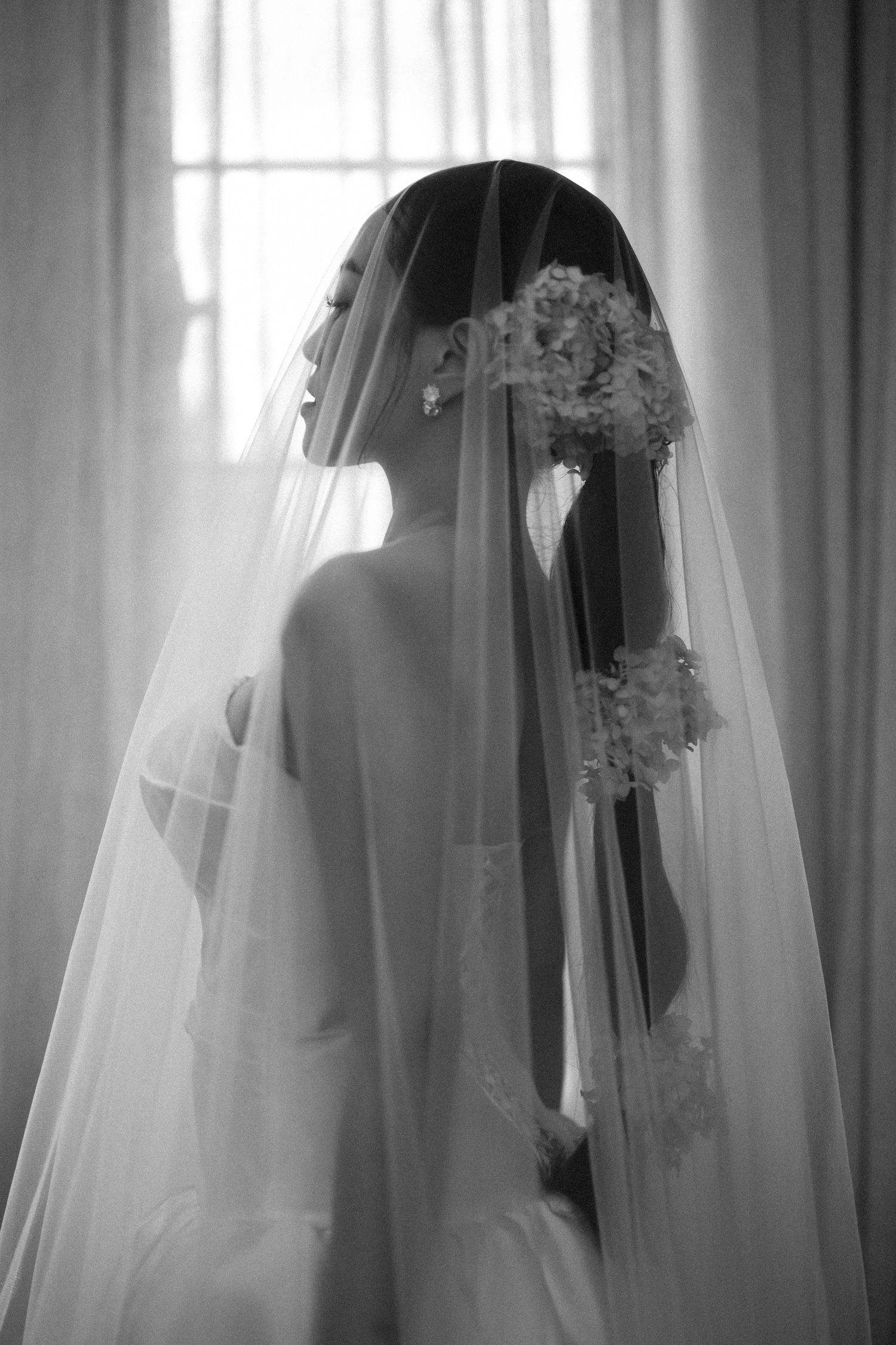 Elegant bride in white wedding gown and flowing veil holding floral bouquet in black and white portrait.