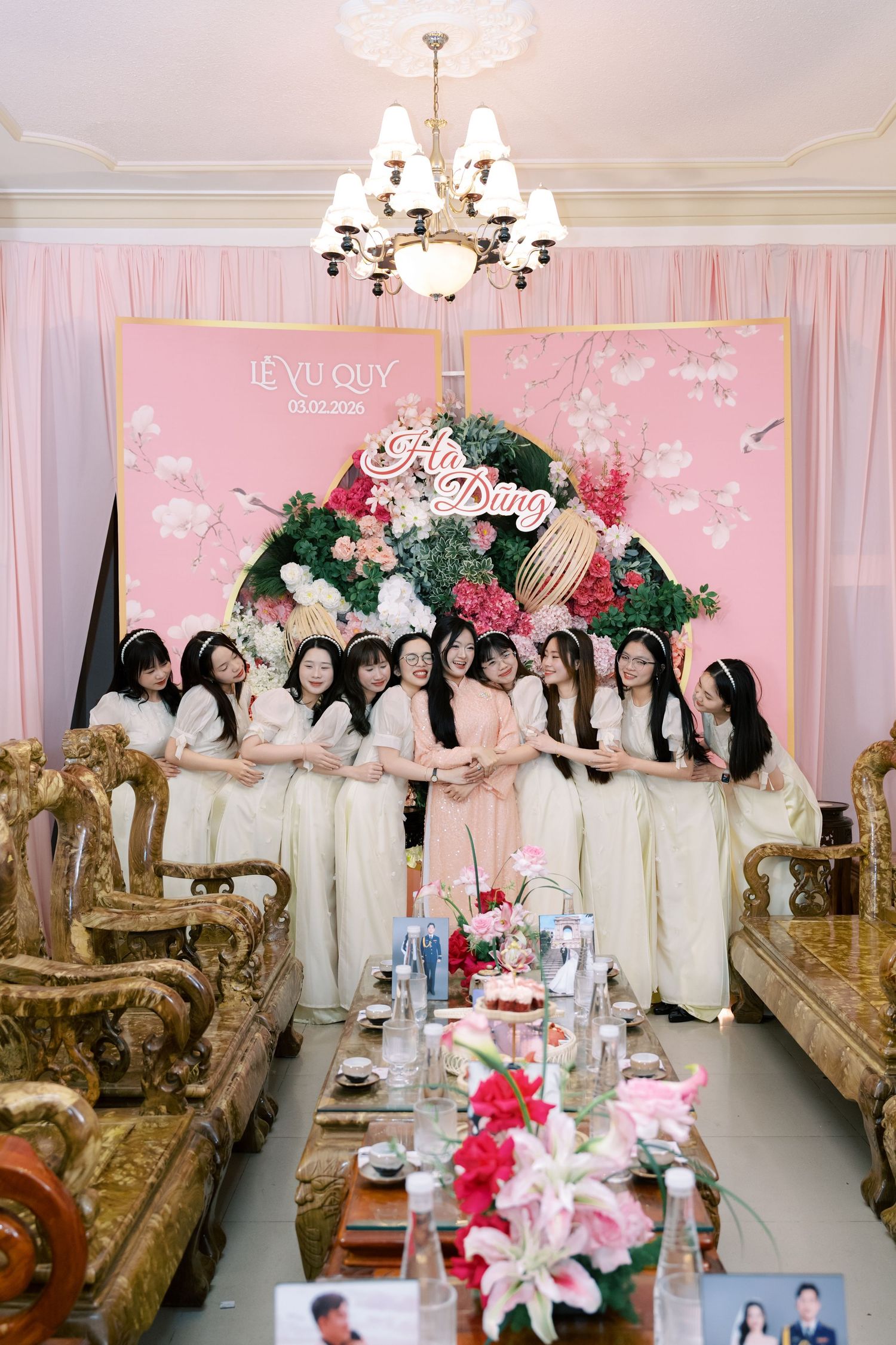Bride in pink ao dai poses with bridesmaids in white dresses before floral wedding backdrop.