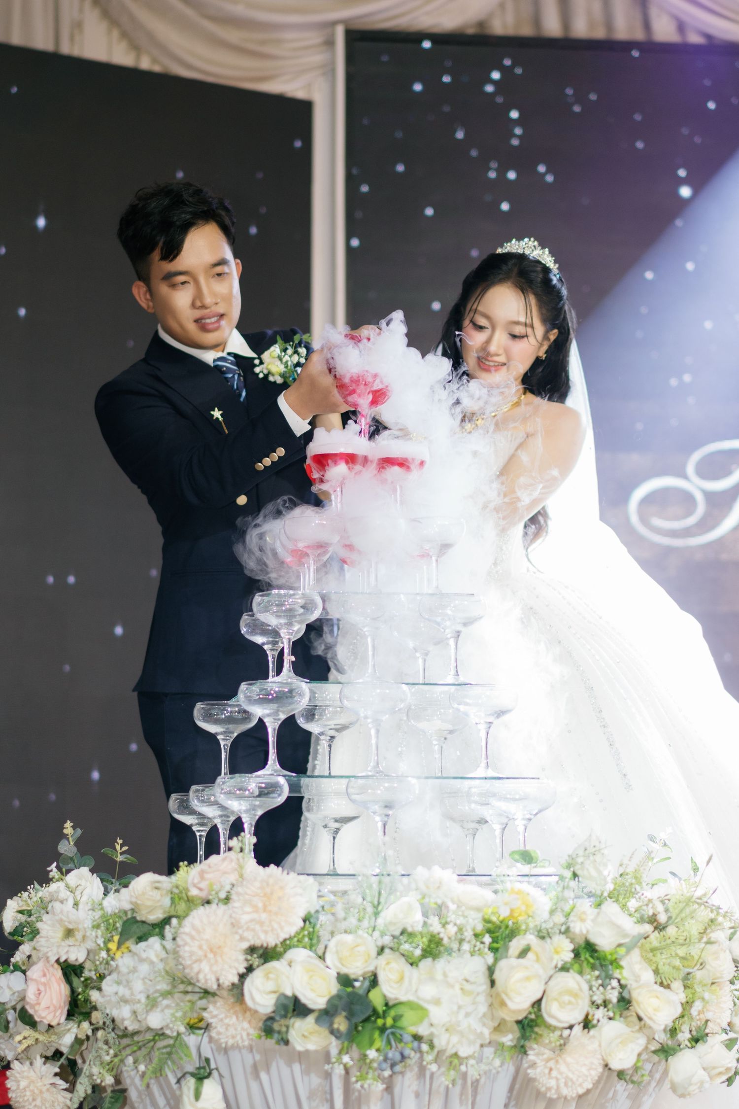 Bride and groom pour champagne tower at elegant wedding reception with smoke effect and white flowers.