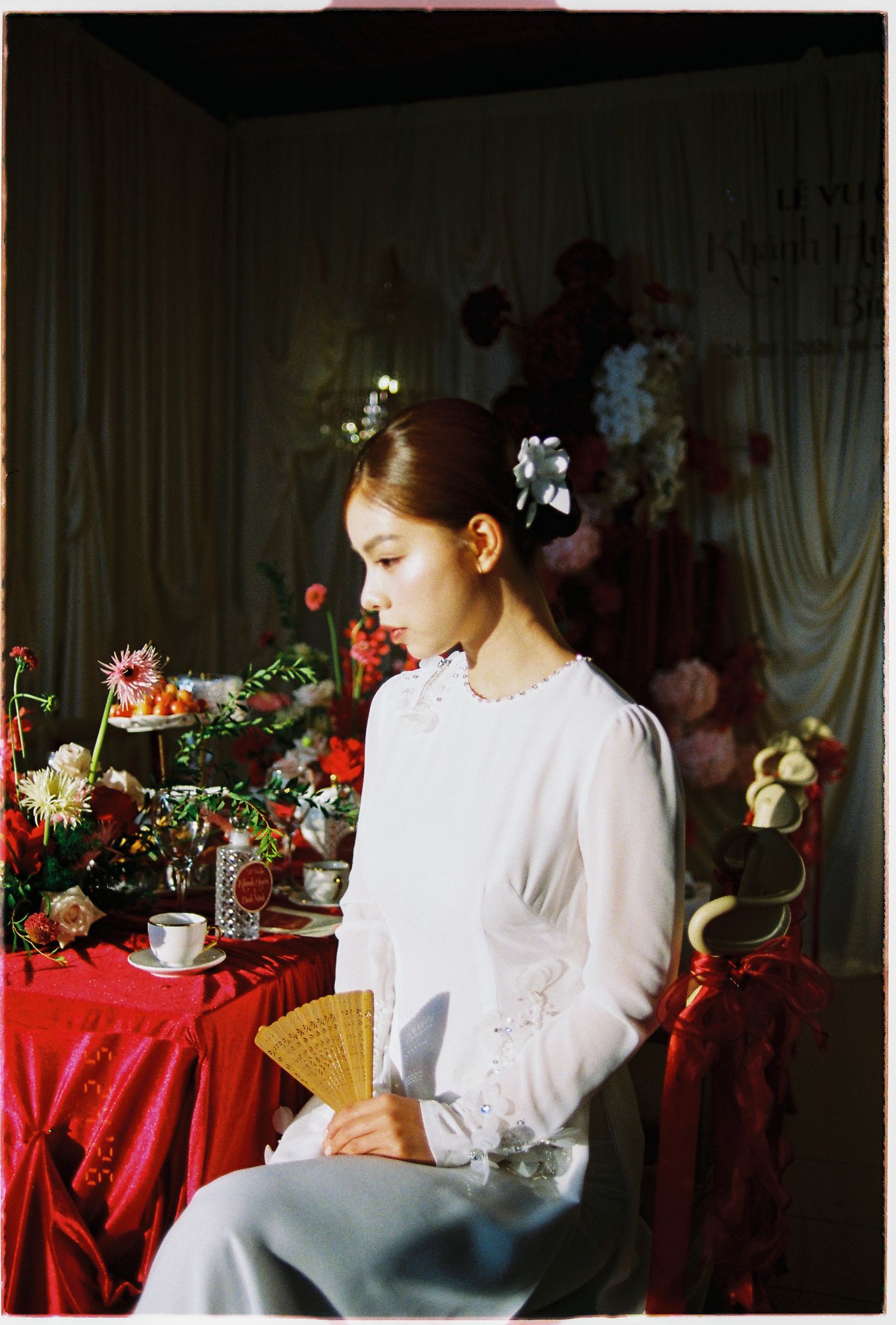 Young woman in white dress holding a fan sits beside red table with flowers in elegant floral studio setting.