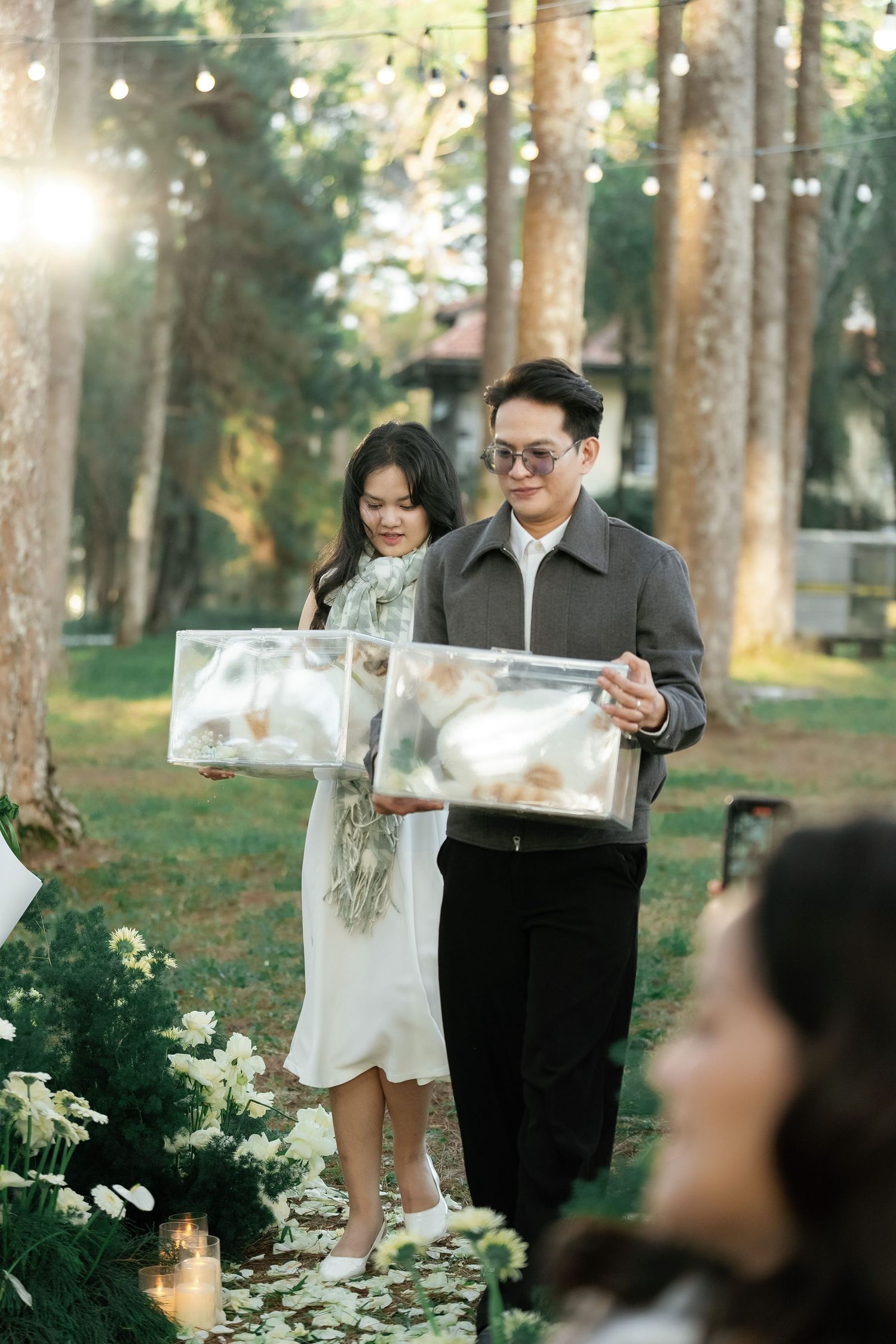 Couple carries clear acrylic boxes with floral decor at an outdoor garden ceremony surrounded by tall trees.