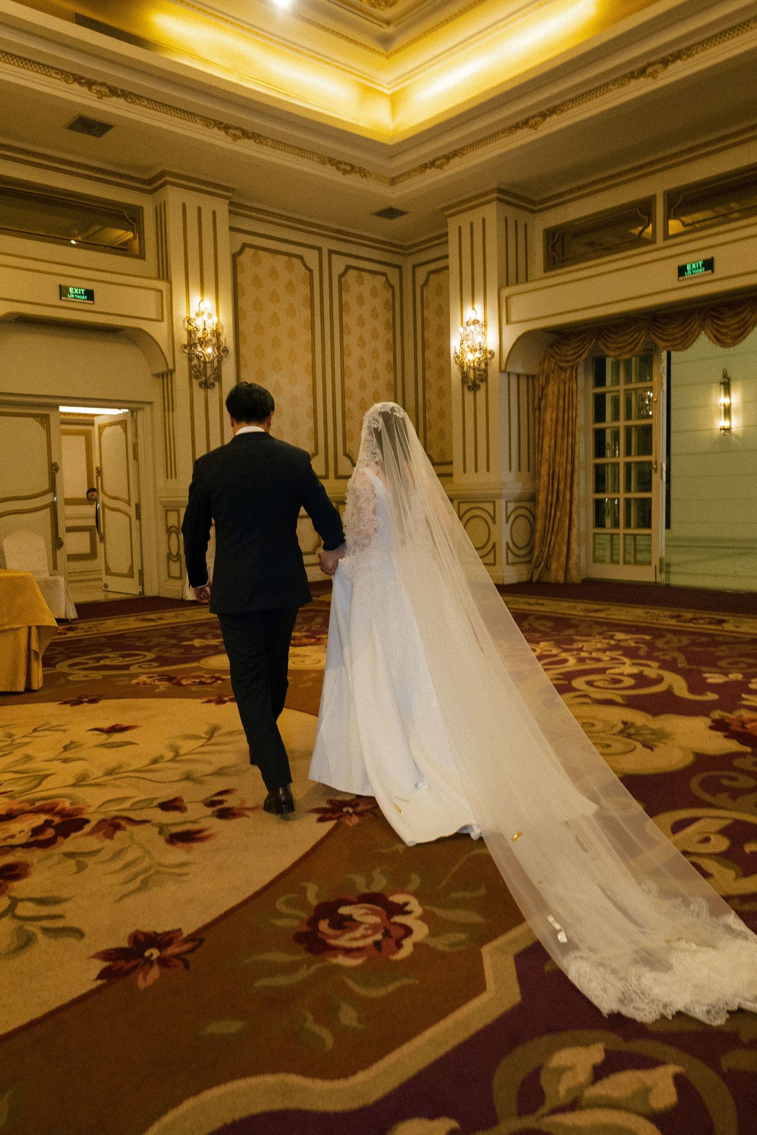 Bride and groom walking hand in hand through an elegant ballroom with ornate decor and floral carpet.