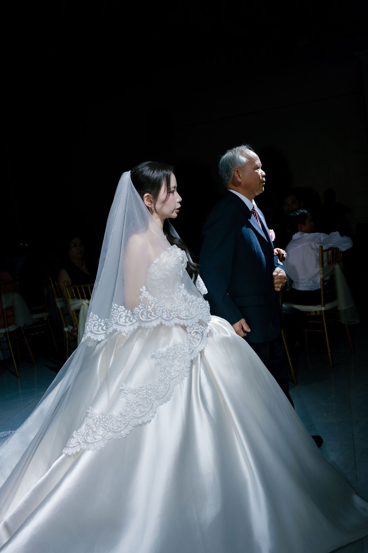 Bride in white lace ballgown wedding dress with veil walks down aisle with older man in dark suit.