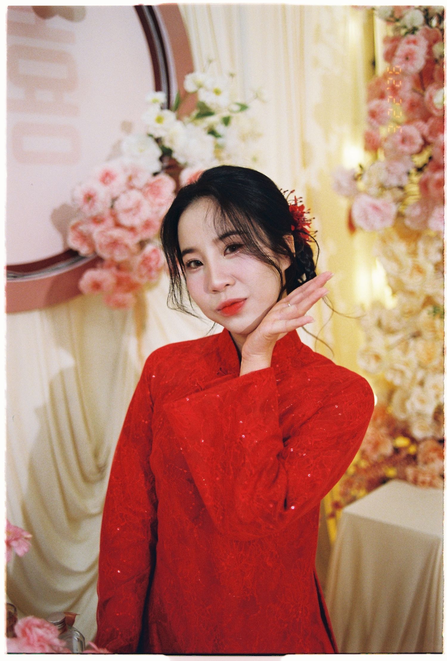 Young woman in red lace ao dai dress posing gracefully among pink floral decorations at a Vietnamese celebration.