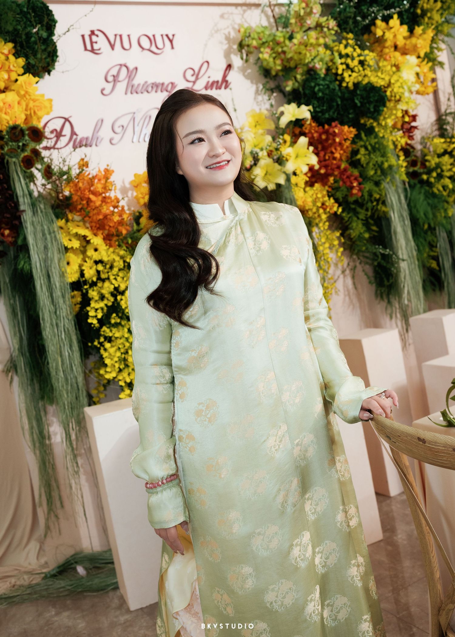 A young woman wearing a light green floral ao dai dress smiles at a Vietnamese engagement ceremony with yellow flowers.
