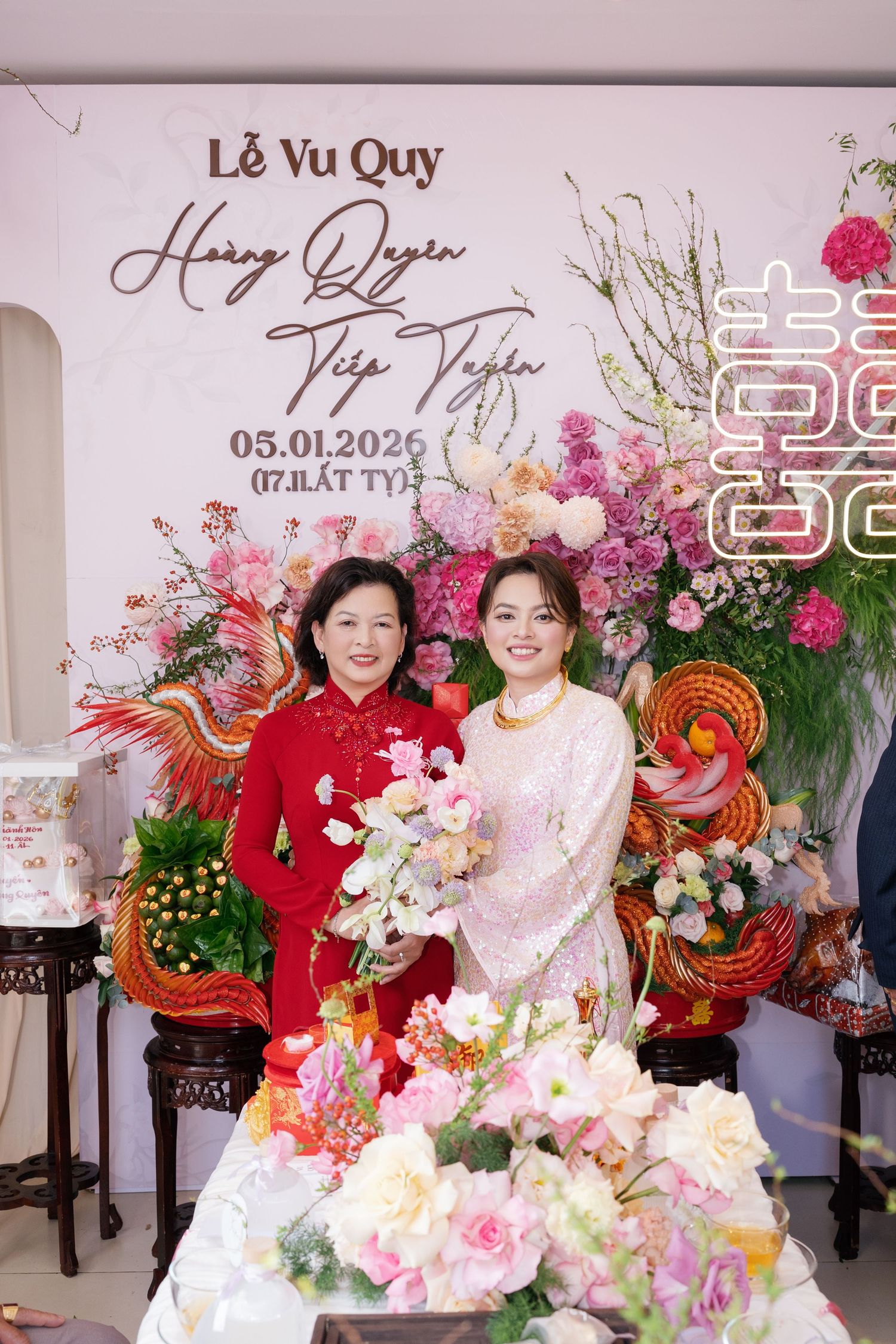 Two women posing together at a Vietnamese wedding ceremony with floral backdrop.