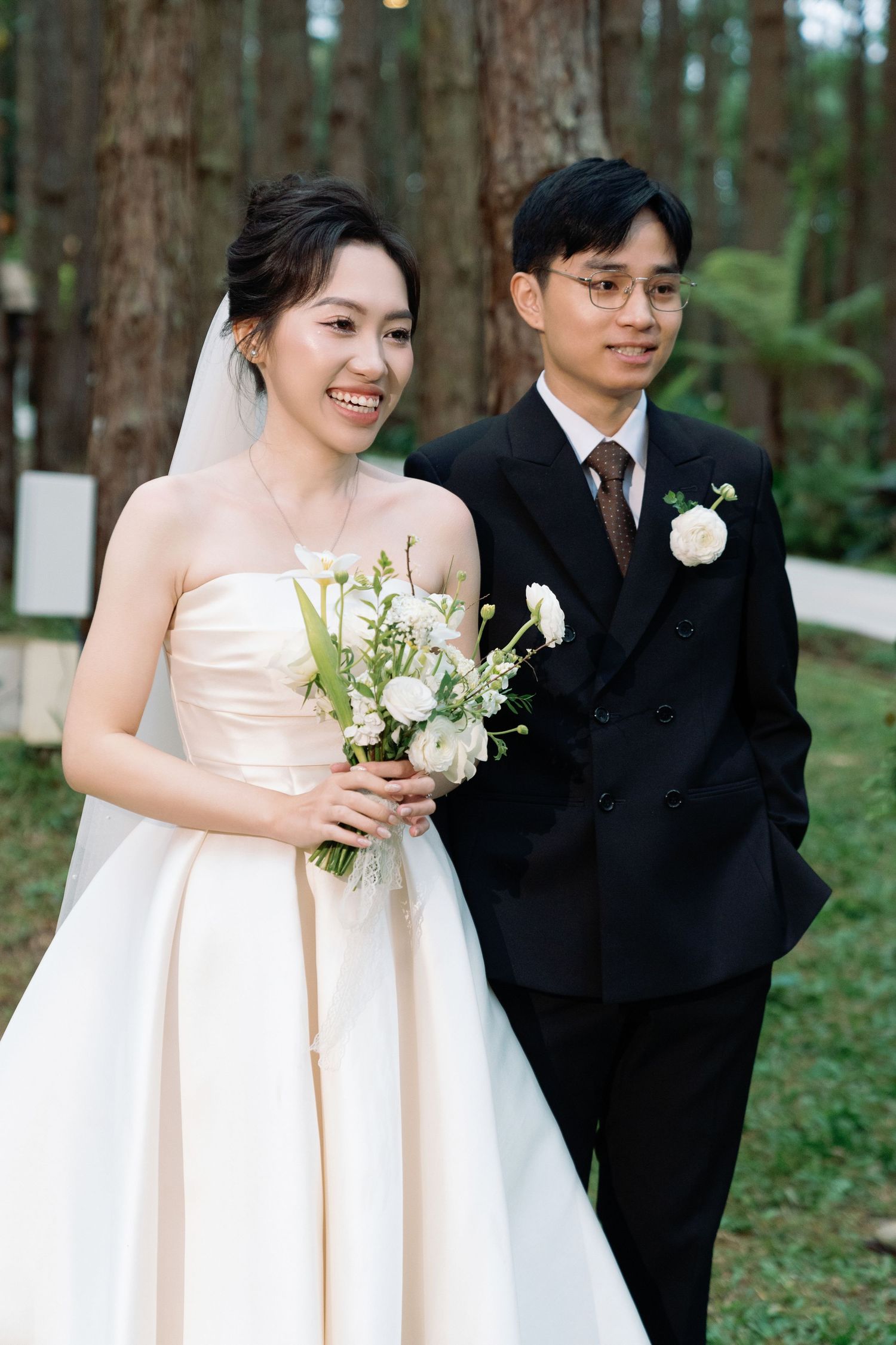 Smiling Asian bride in strapless white gown holding bouquet stands with groom in black suit at outdoor forest wedding.