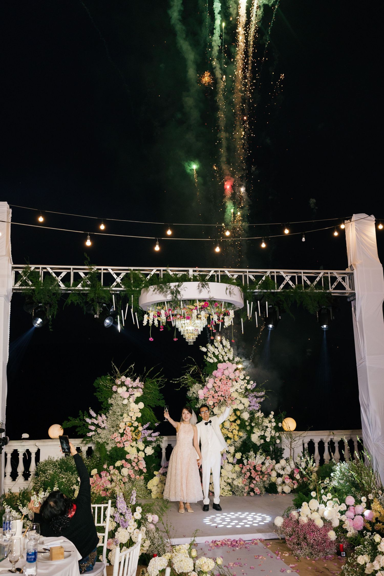Bride and groom celebrate under fireworks at an outdoor night wedding with floral arrangements and string lights.