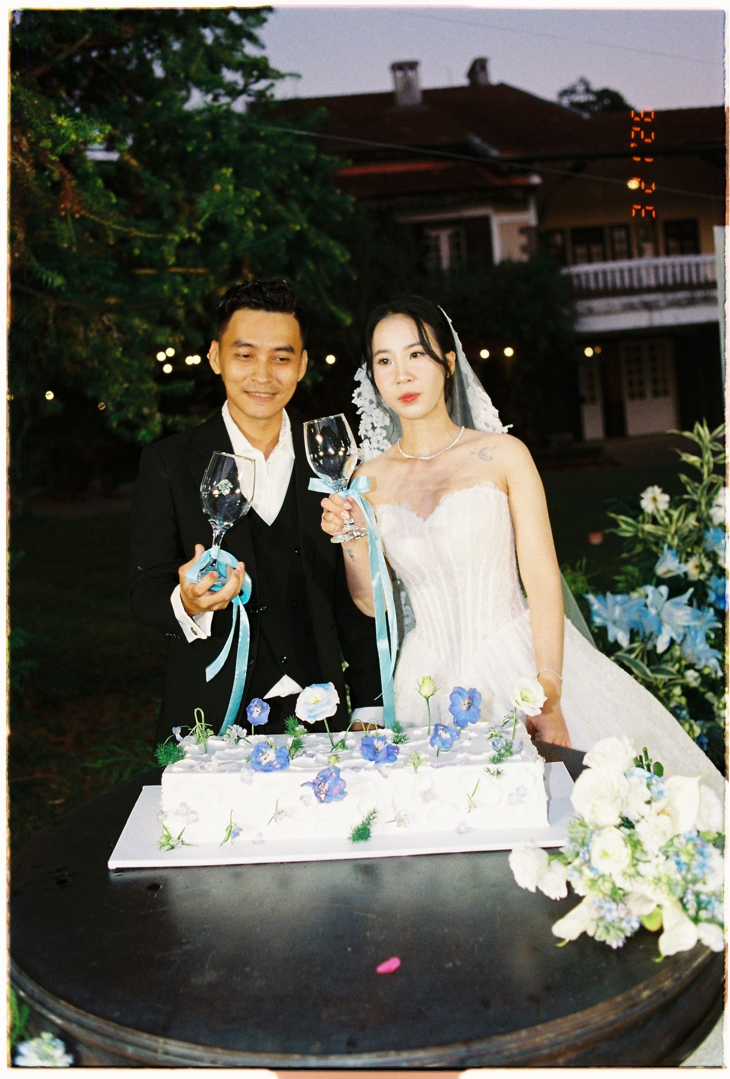 Bride and groom toasting with wine glasses beside a white wedding cake decorated with blue flowers at an outdoor evening reception.