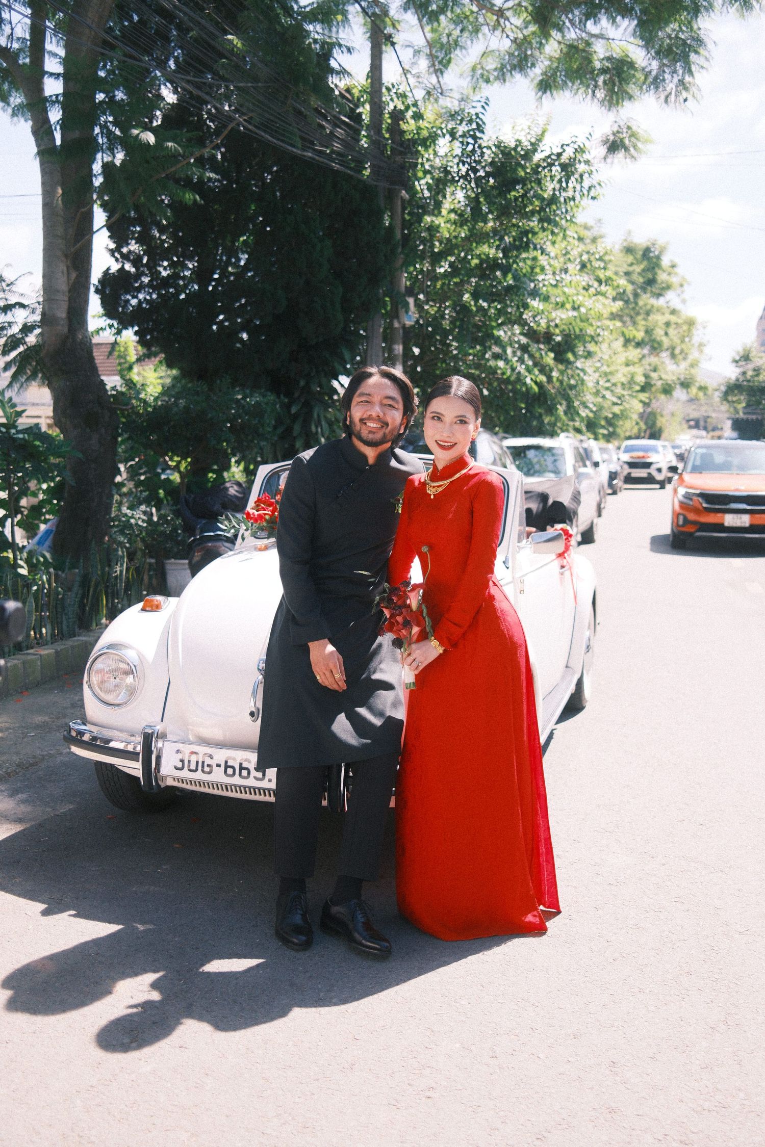 Couple in formal attire posing by a white vintage Volkswagen Beetle on a sunny street, woman in red ao dai gown.