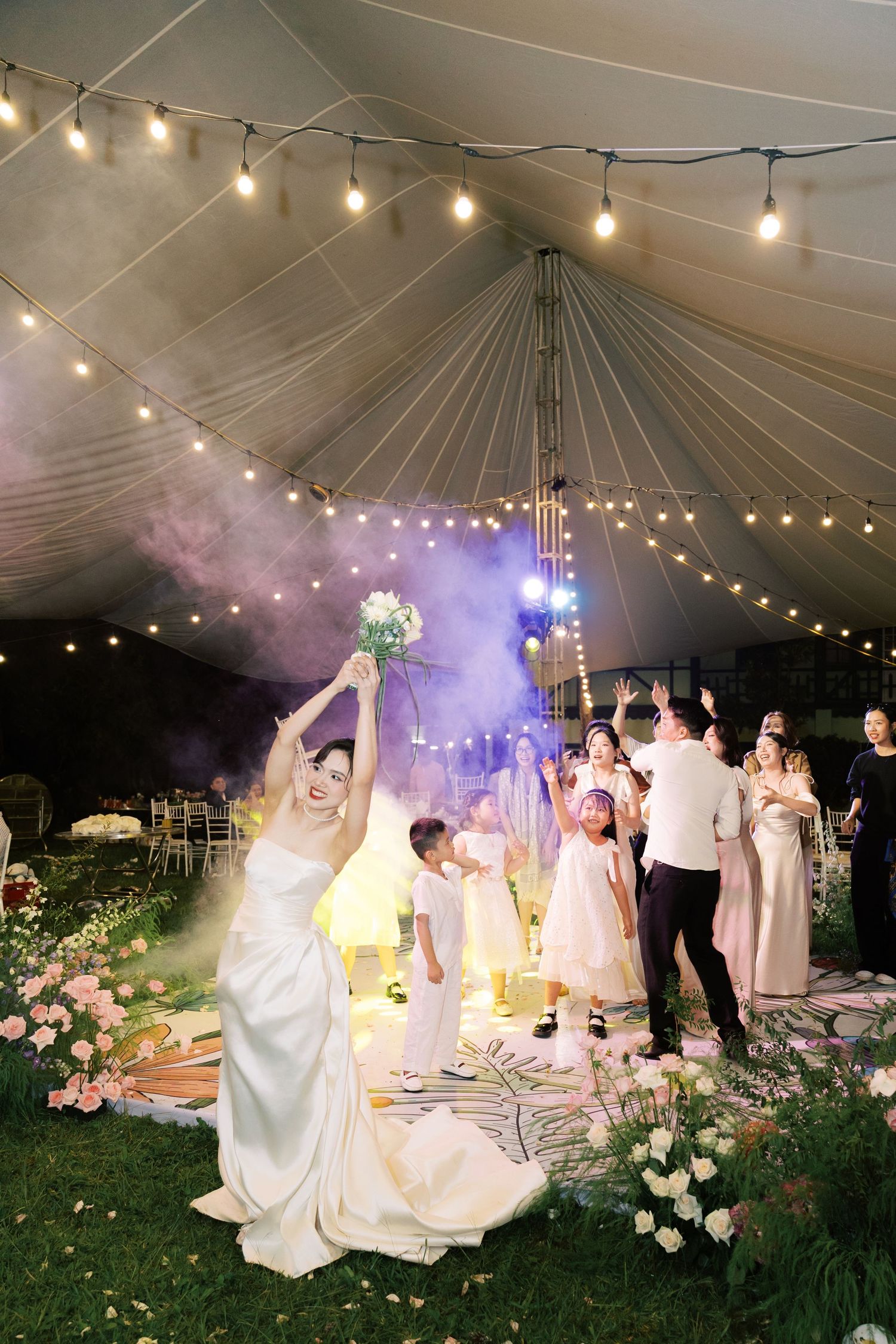 Bride tosses bouquet under glowing string lights at elegant outdoor wedding reception tent at night.