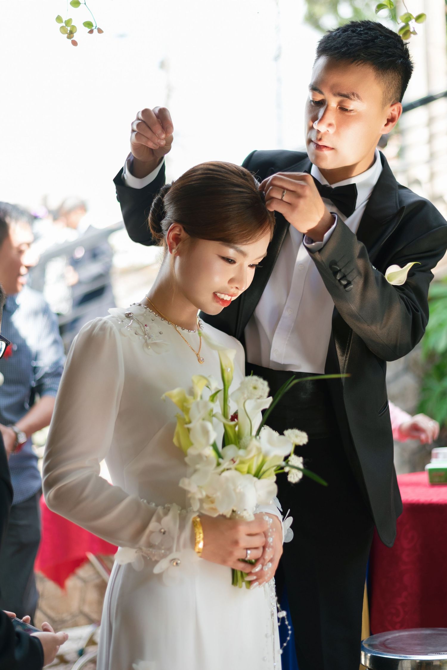 Groom in black tuxedo placing hairpin on bride in white ao dai holding calla lily bouquet at traditional wedding ceremony.