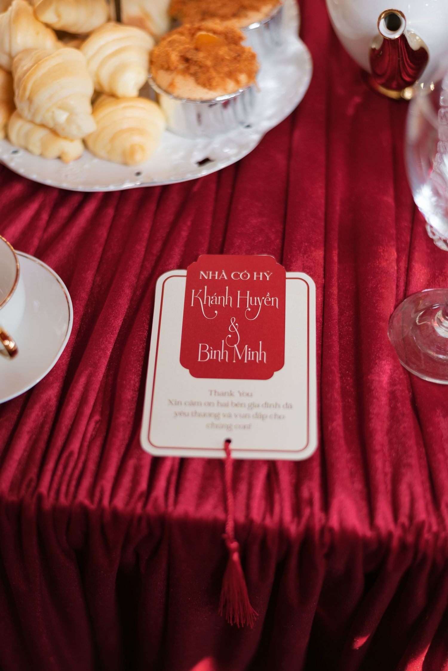 Elegant red velvet table with a Vietnamese wedding card for Khanh Huyen and Binh Minh, beside pastries and glassware.