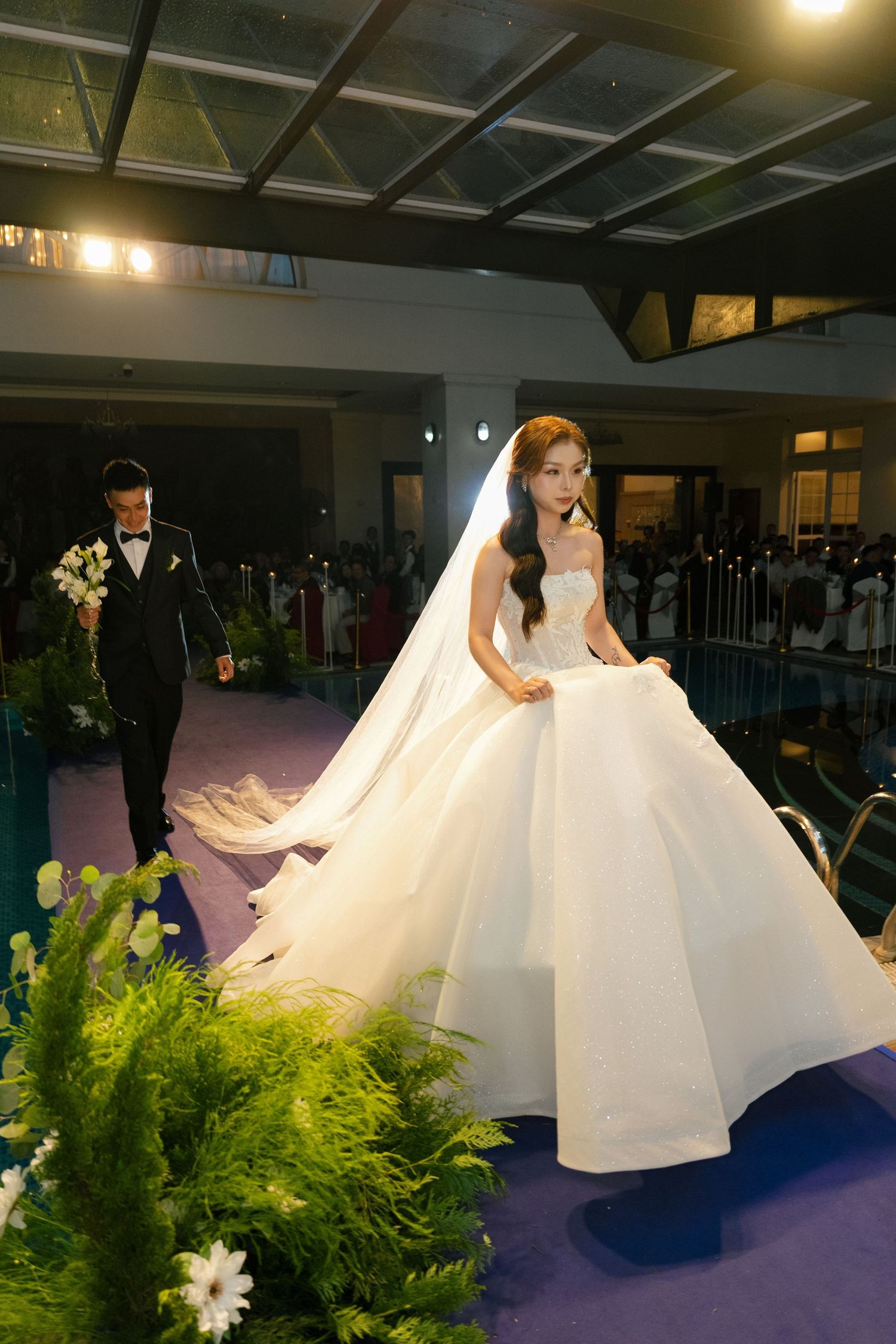 Bride in a stunning white ballgown with long veil walks the aisle as groom follows at an elegant wedding ceremony.