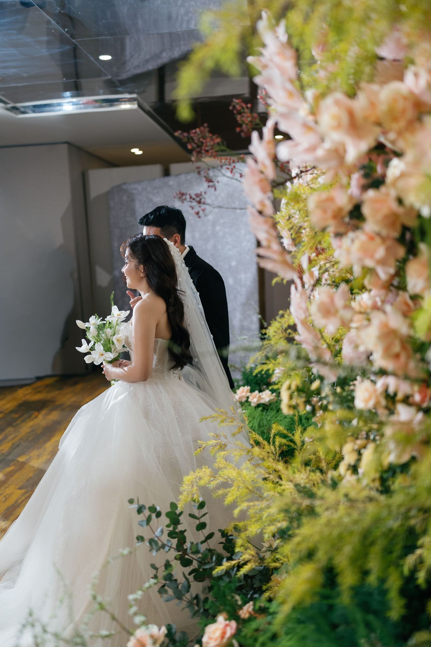Bride in white ball gown and groom stand among lush greenery and blush flowers at wedding.