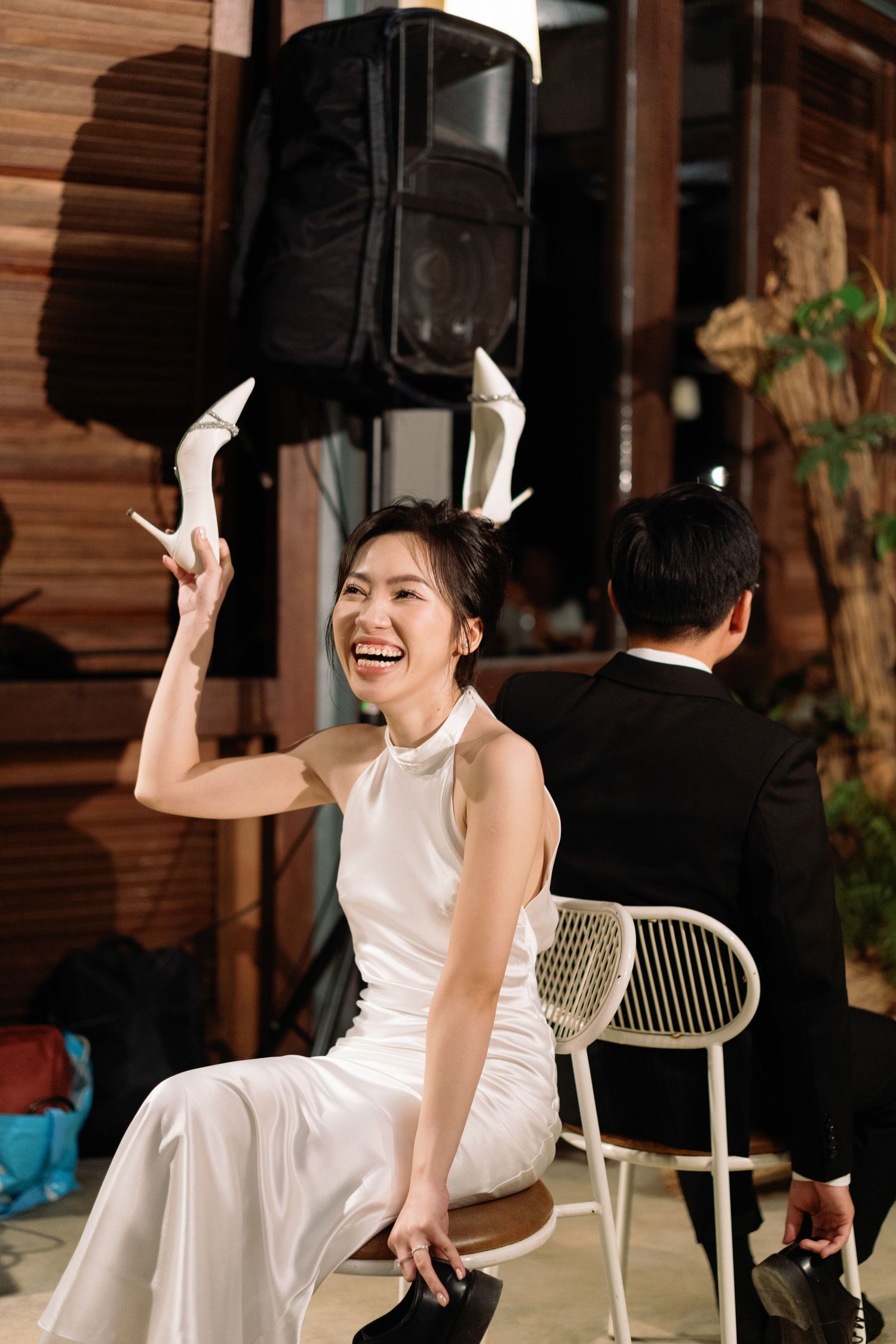 Joyful Asian bride in white halter dress laughing while holding up white heels at wedding reception.
