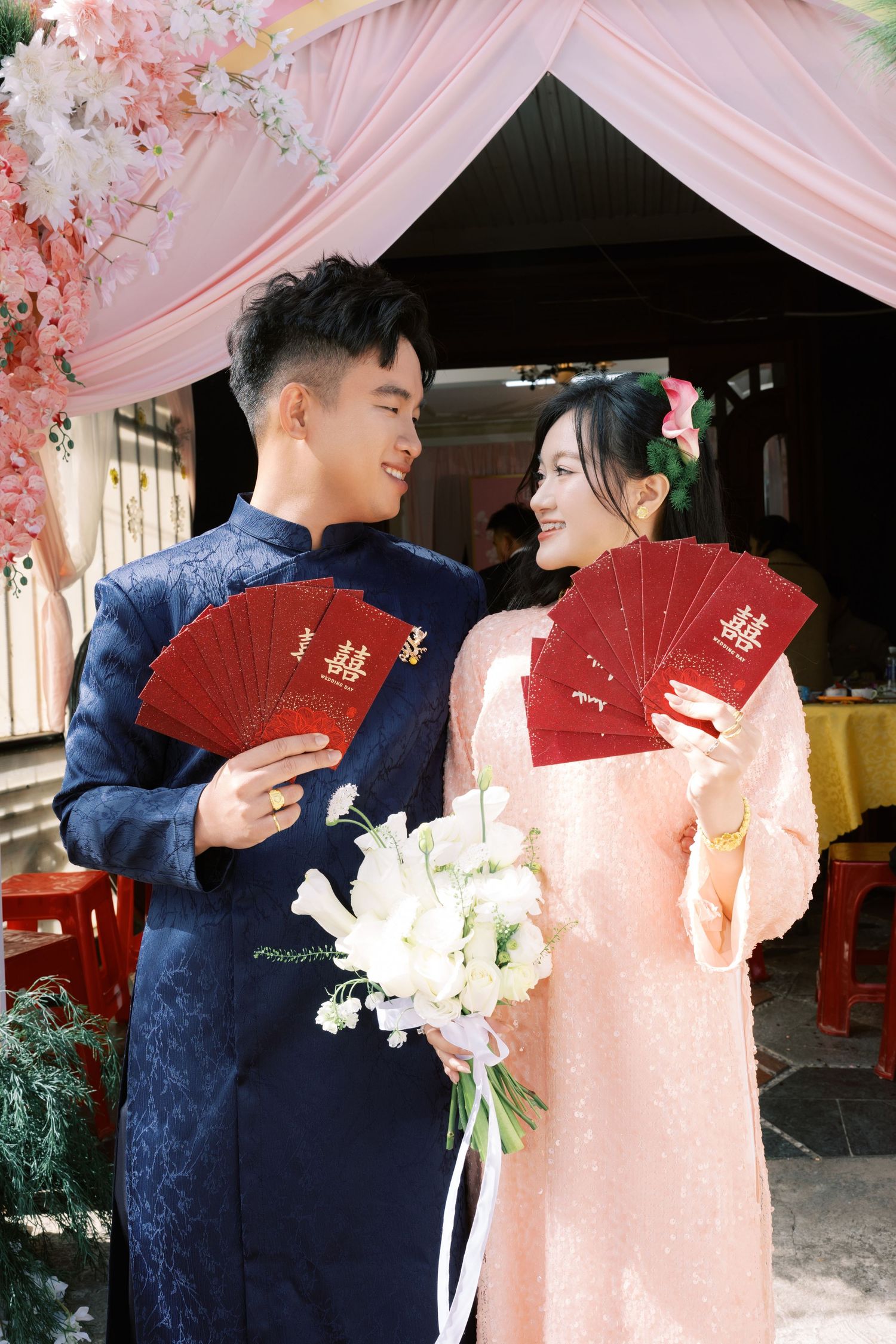 Vietnamese couple in traditional ao dai attire holding red envelopes and white flowers at a wedding ceremony.