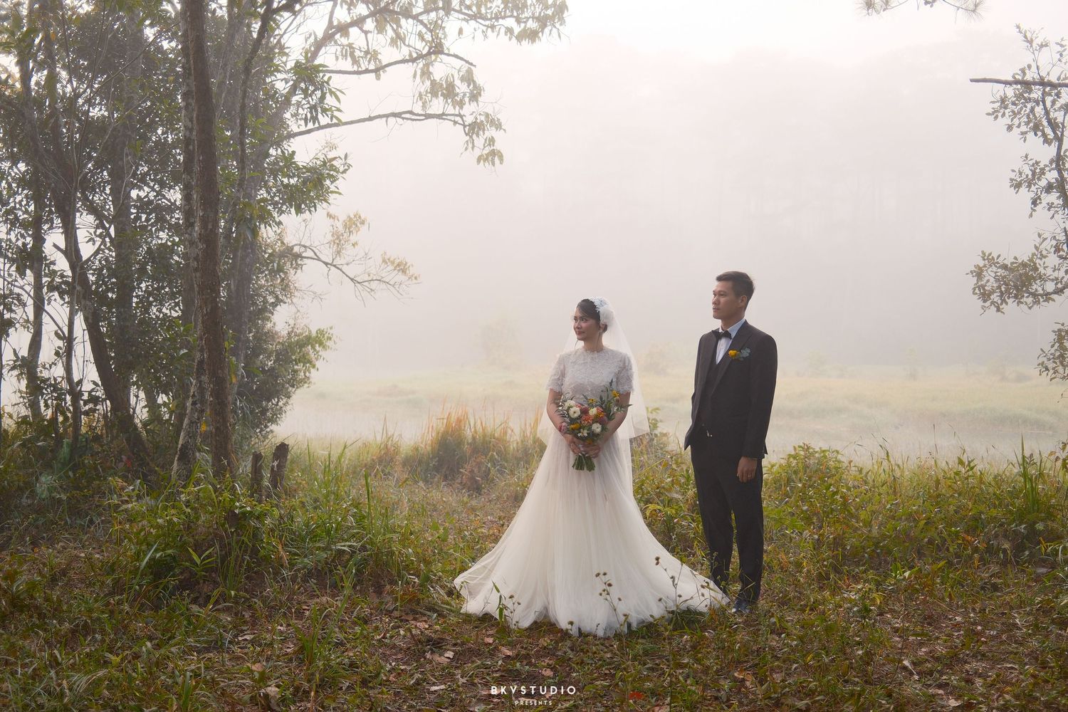 Bride and groom stand apart in misty forest clearing, she in white gown with bouquet, he in black suit.