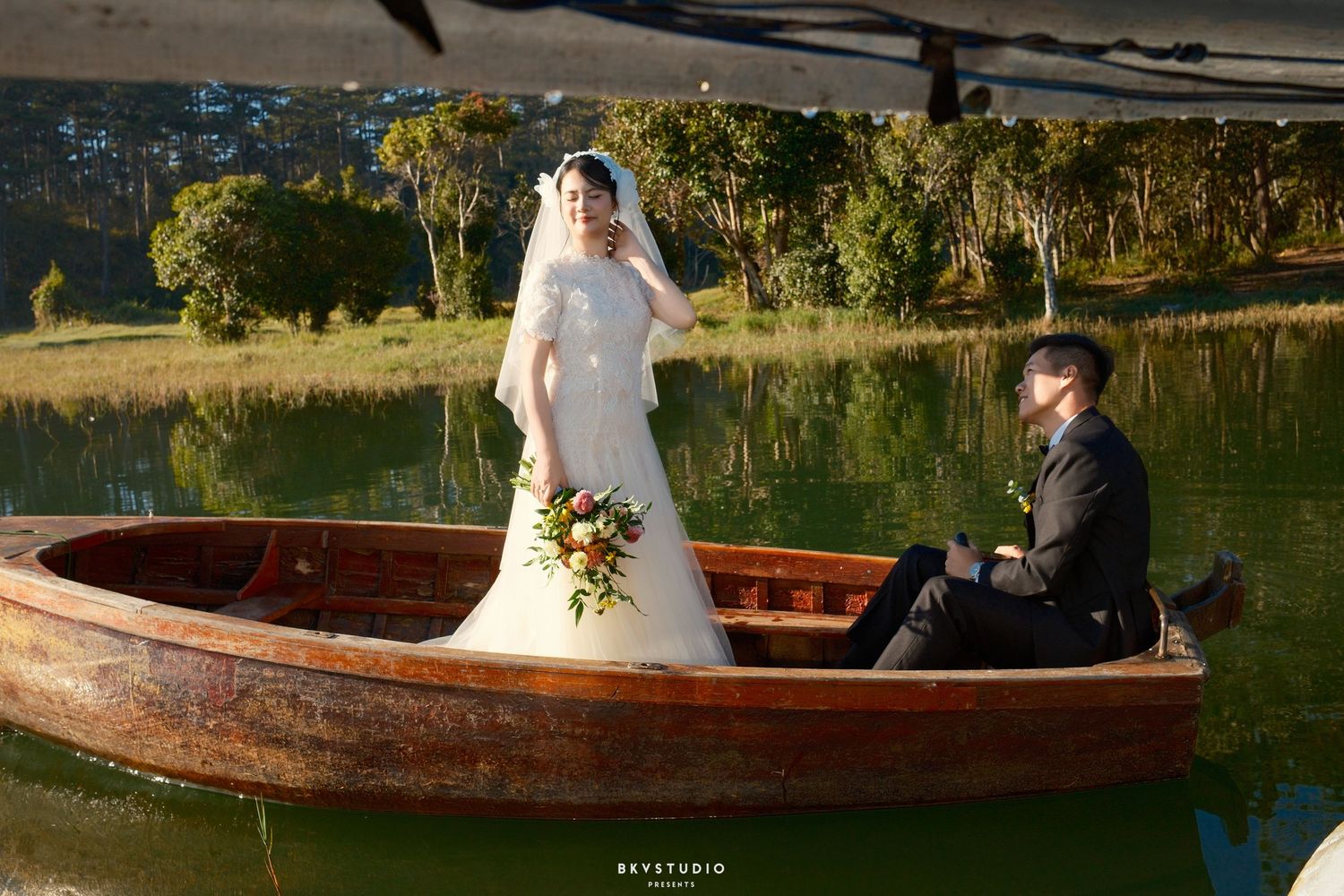 Bride stands holding bouquet as groom sits in wooden boat on serene lake during wedding photography.