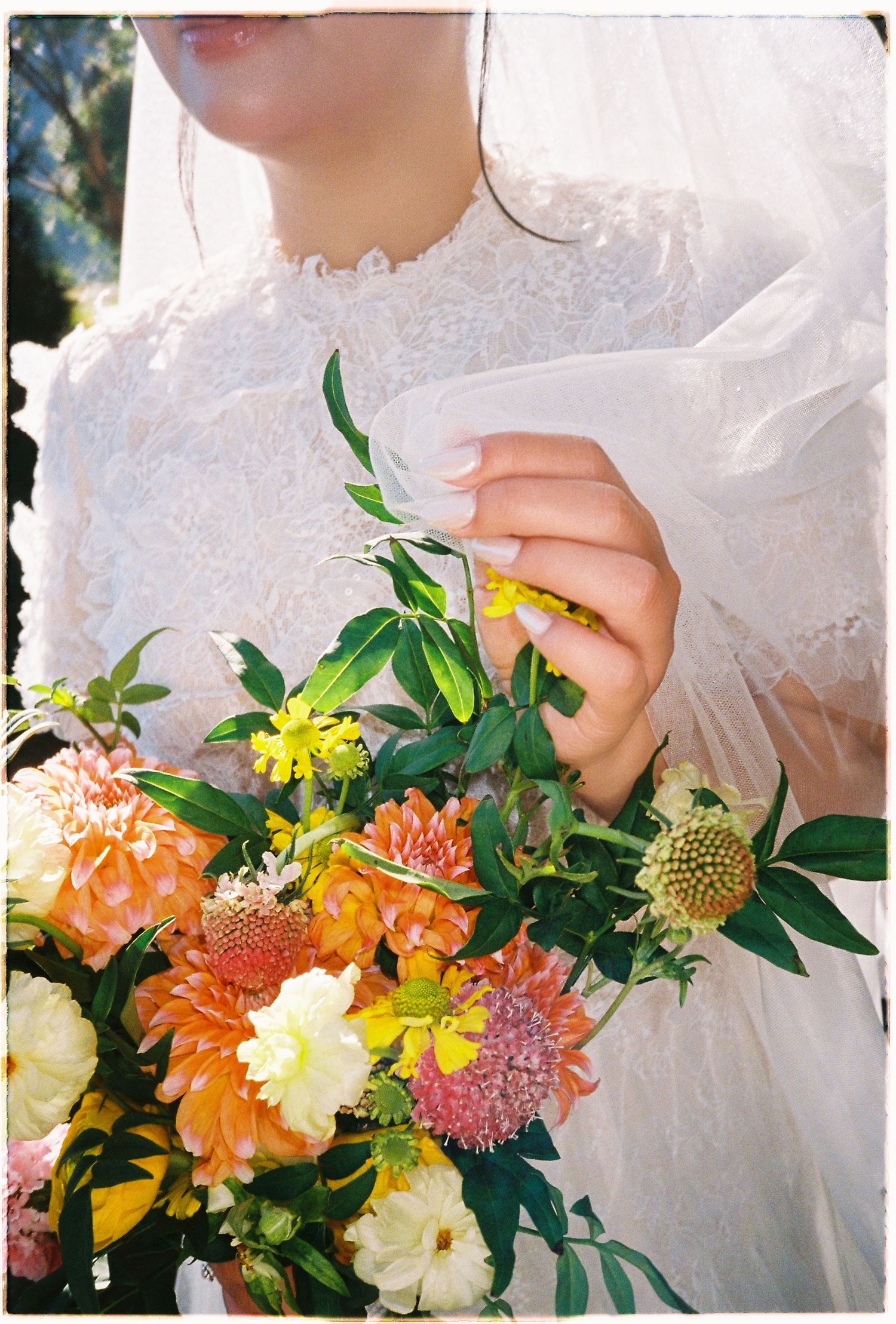 Close-up of bride in white lace dress holding vibrant orange and yellow wedding bouquet outdoors.