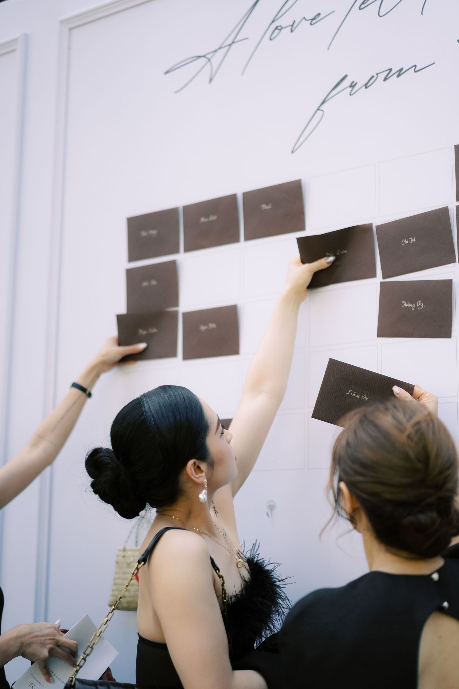 Wedding guests placing dark envelope seating cards on a white wall at a reception venue.
