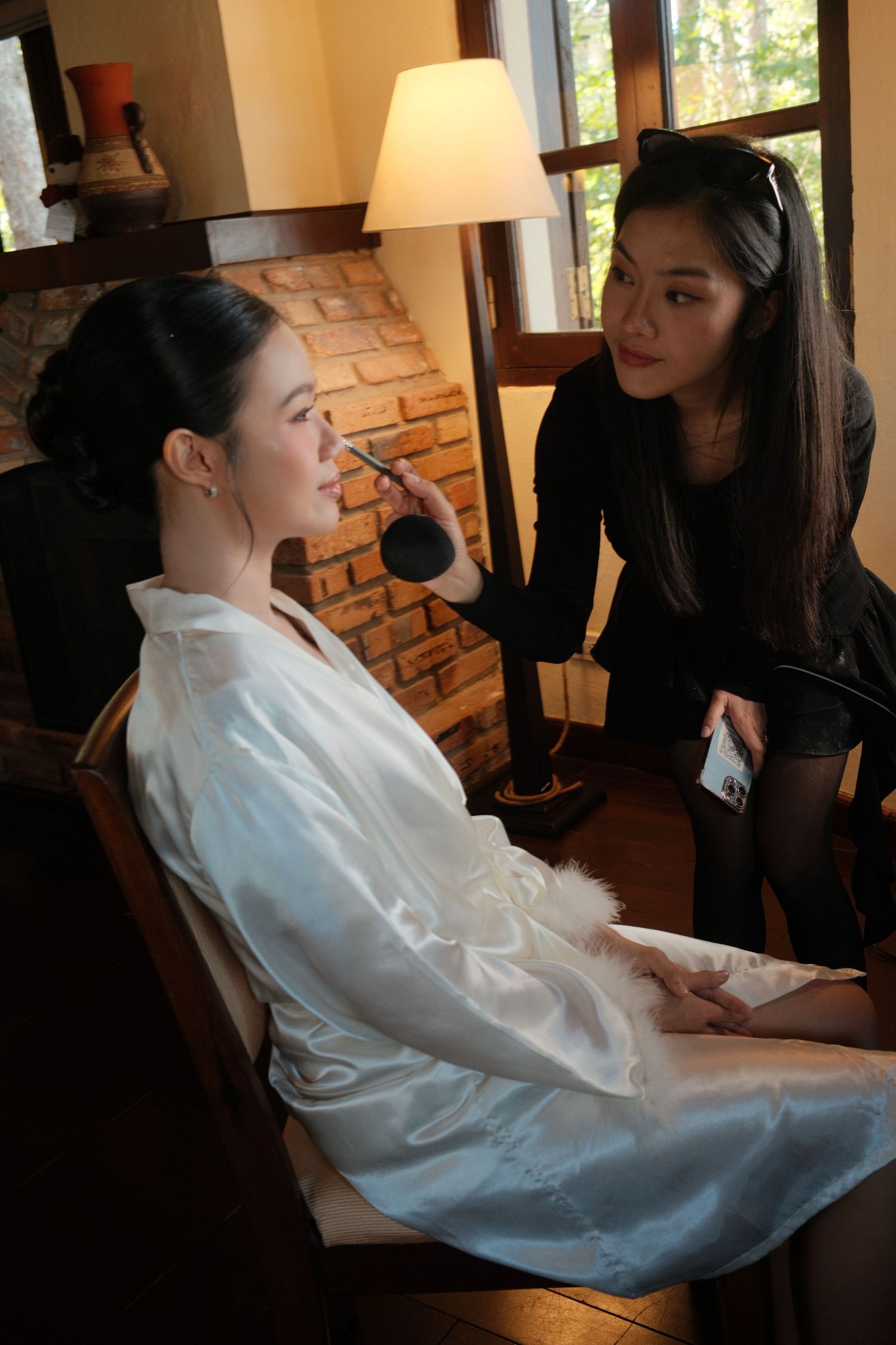 A makeup artist applies cosmetics to a bride in a white robe seated in a warmly lit rustic room.
