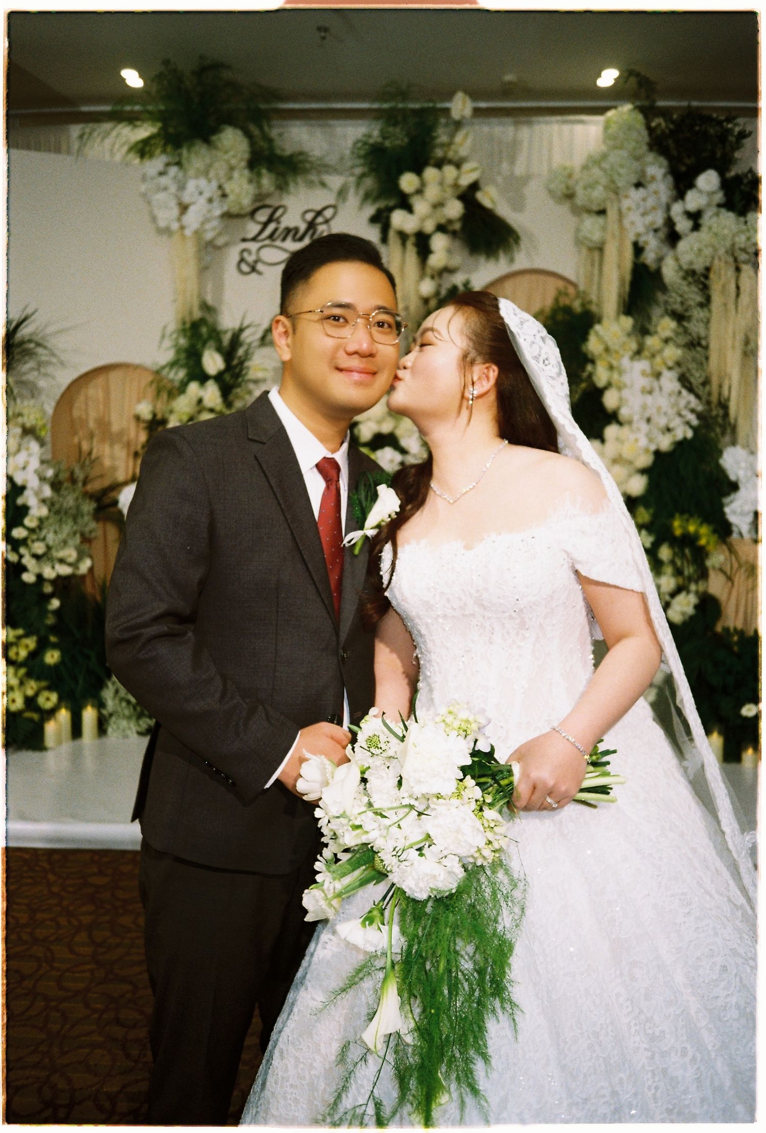Bride kissing groom on cheek at wedding altar decorated with white floral arrangements, film photo.