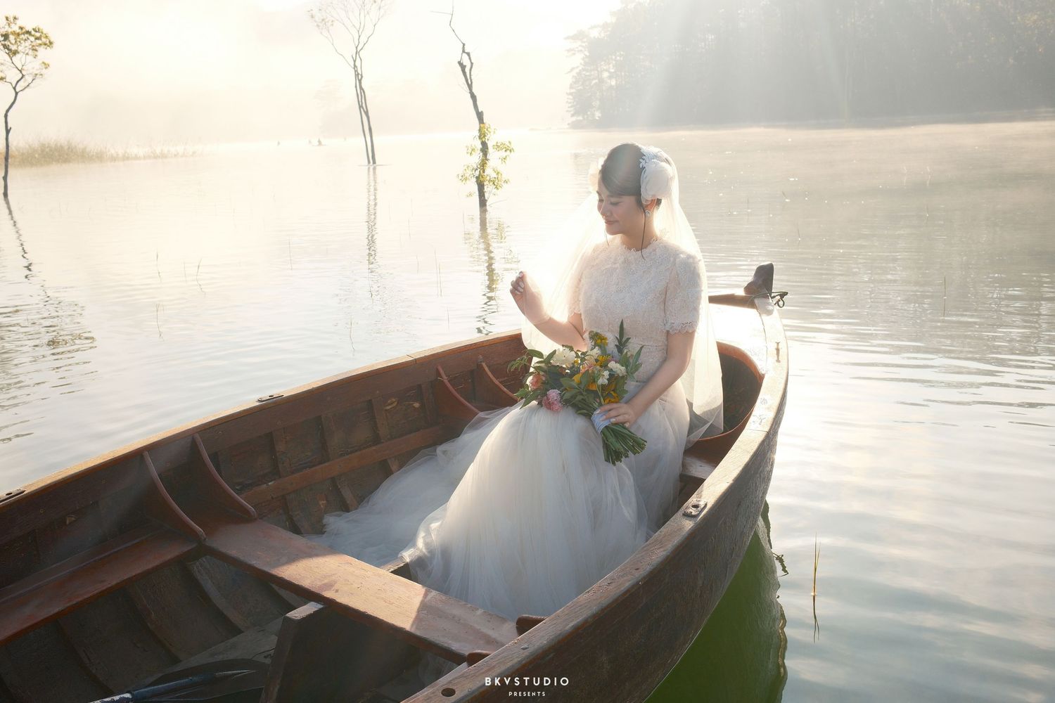 Bride in white lace gown sits alone in wooden boat on serene misty lake holding bouquet.