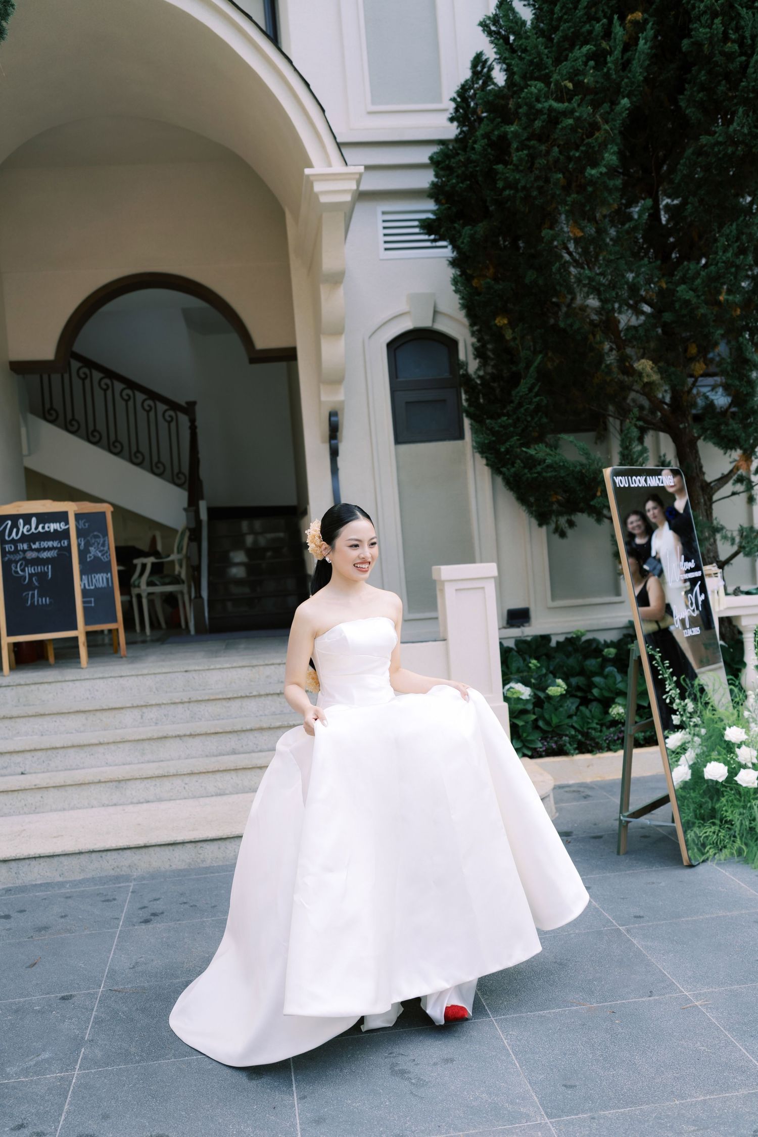 Smiling bride in strapless white ball gown with red heels walking outside elegant venue.