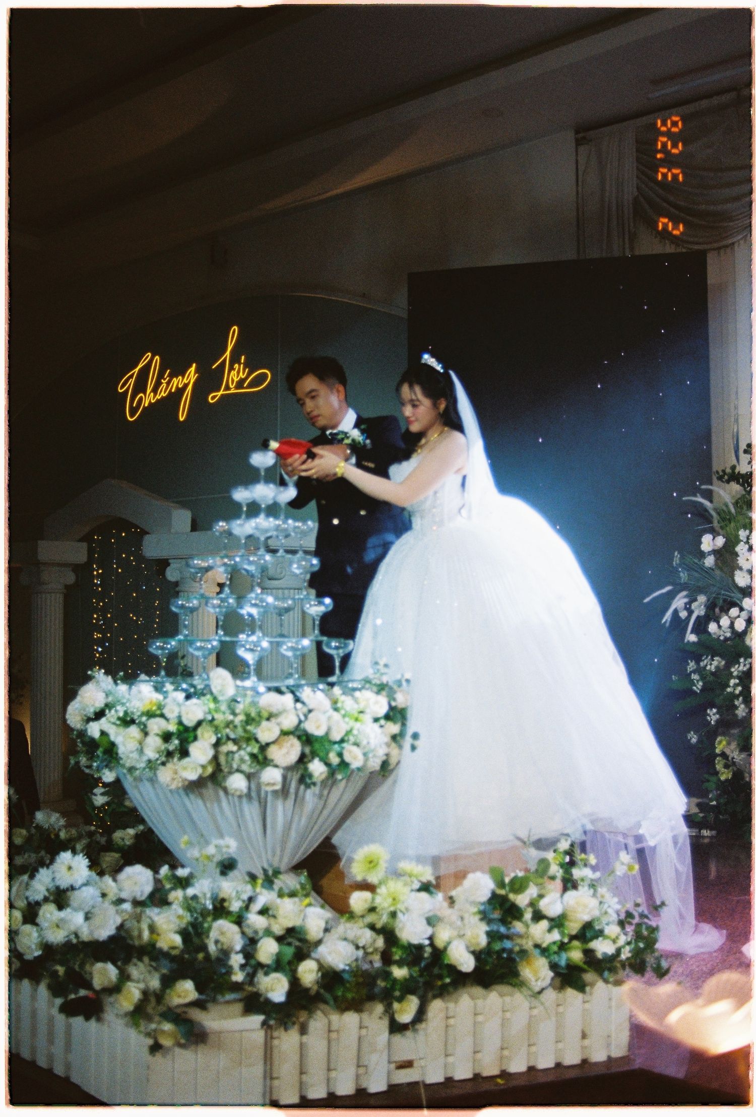 Bride and groom pour champagne tower at floral-decorated wedding reception with neon sign backdrop.
