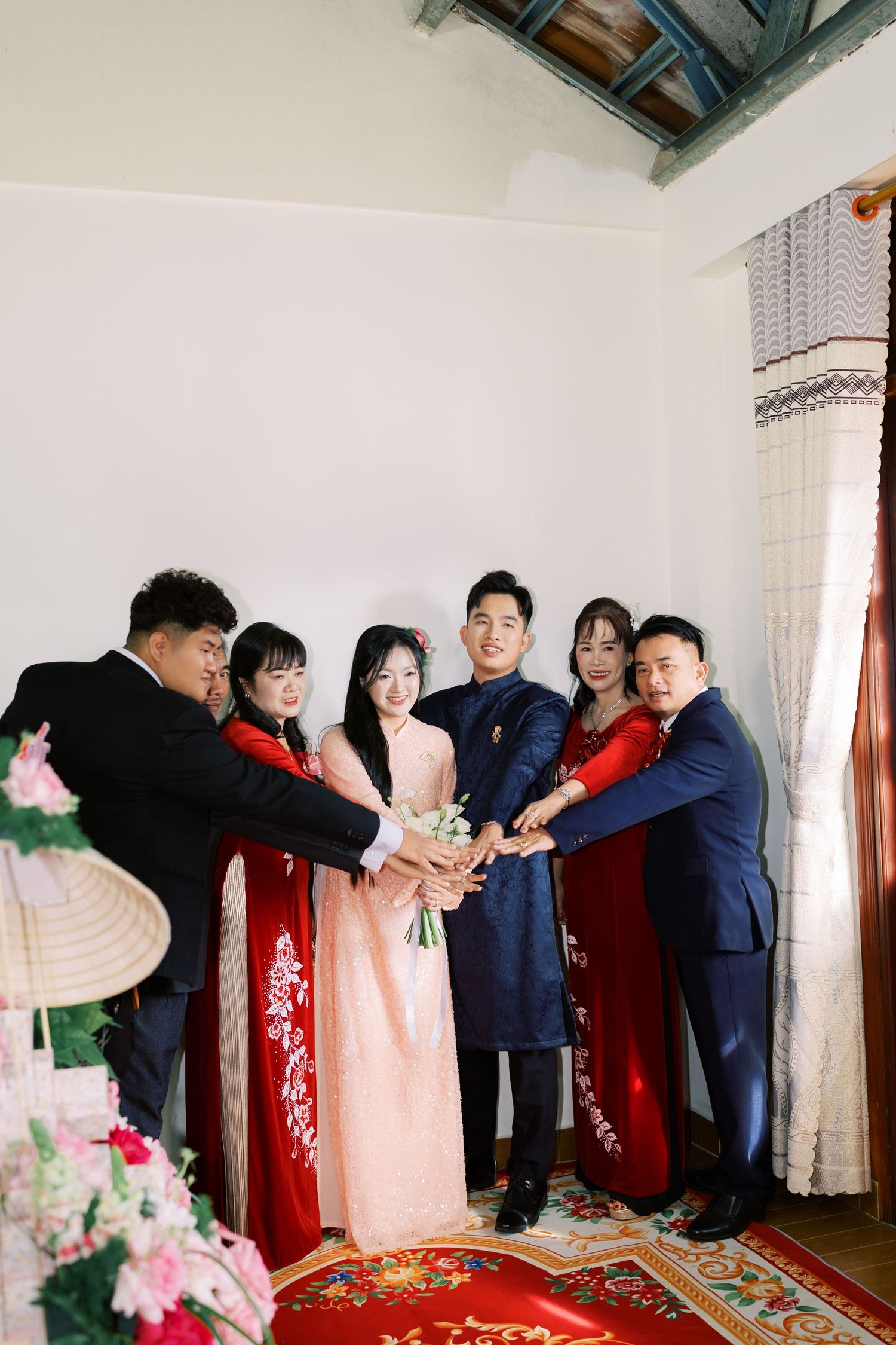 Vietnamese wedding party in traditional ao dai and suits joins hands over bridal bouquet indoors.