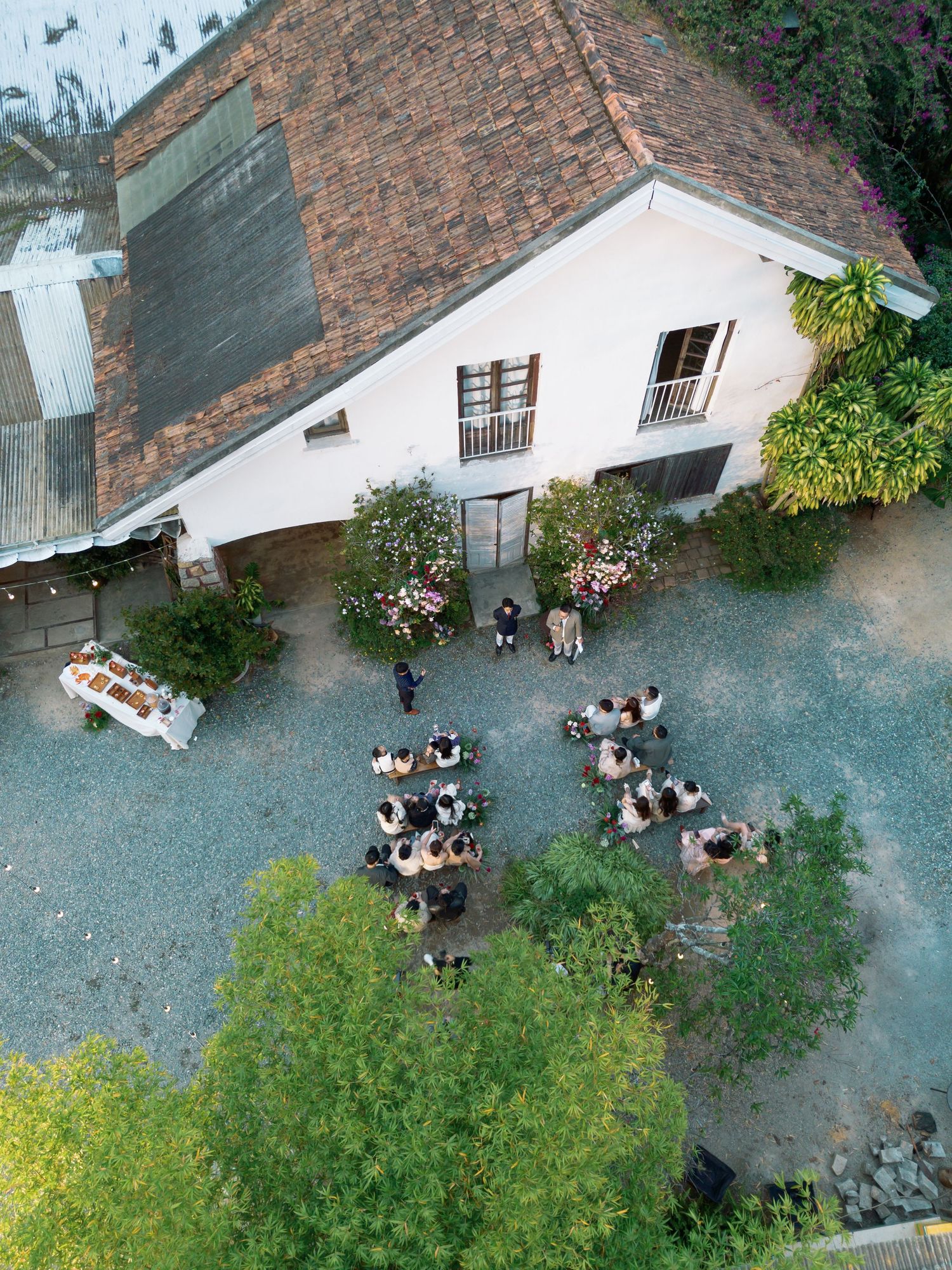 Aerial view of an outdoor wedding ceremony in a rustic courtyard beside a white barn with a terracotta roof.
