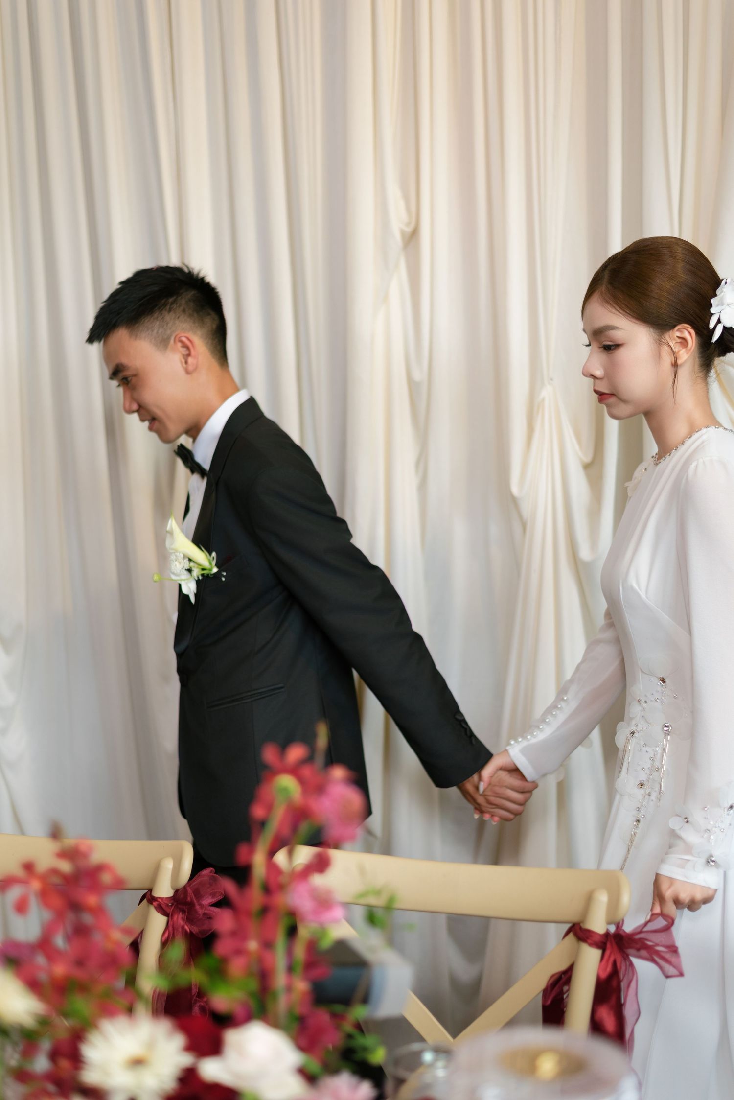 Asian couple in wedding attire holding hands at reception table, groom in black tuxedo, bride in white gown with floral decor.