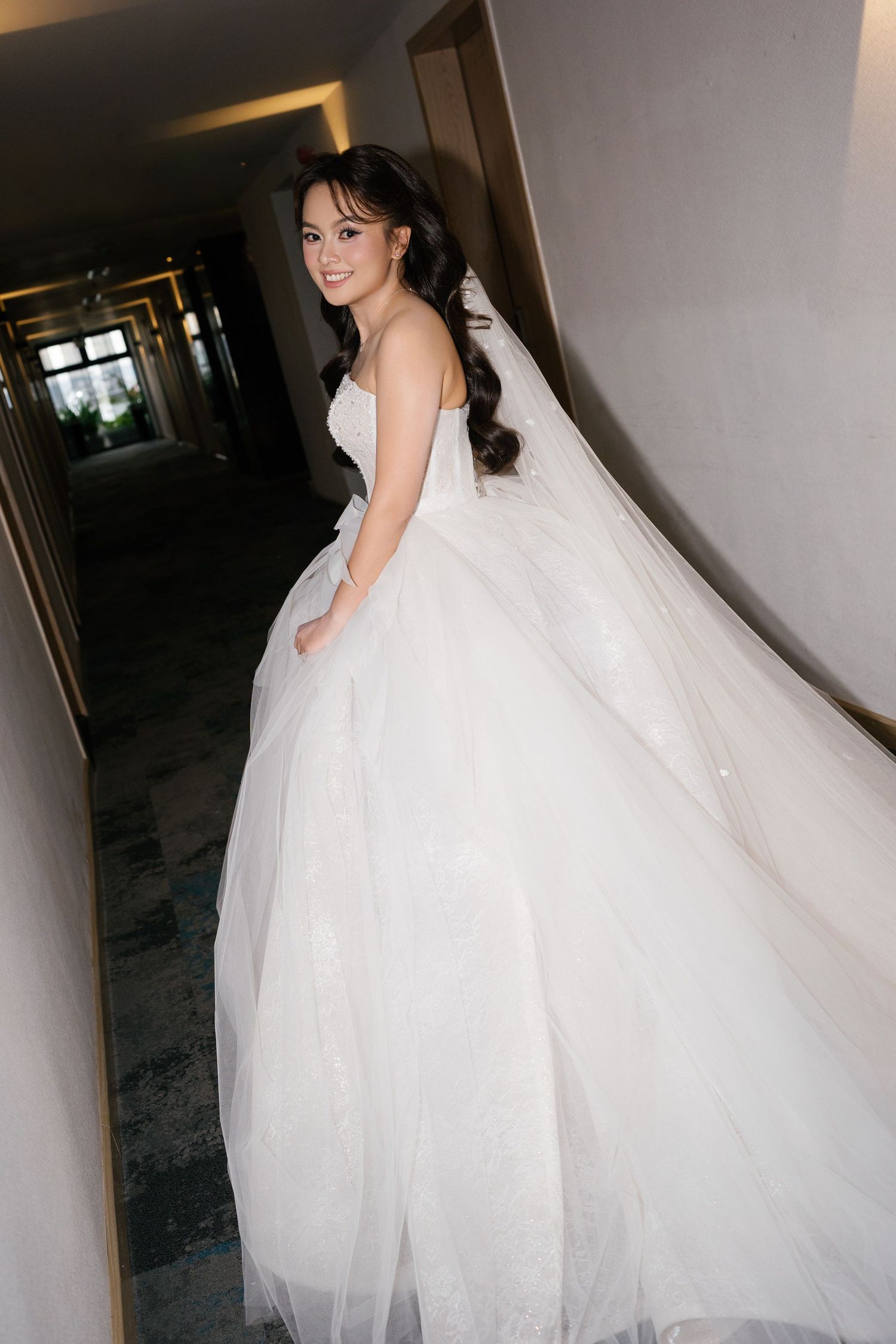 A bride in a flowing white strapless ball gown with a long veil poses in a hotel corridor.